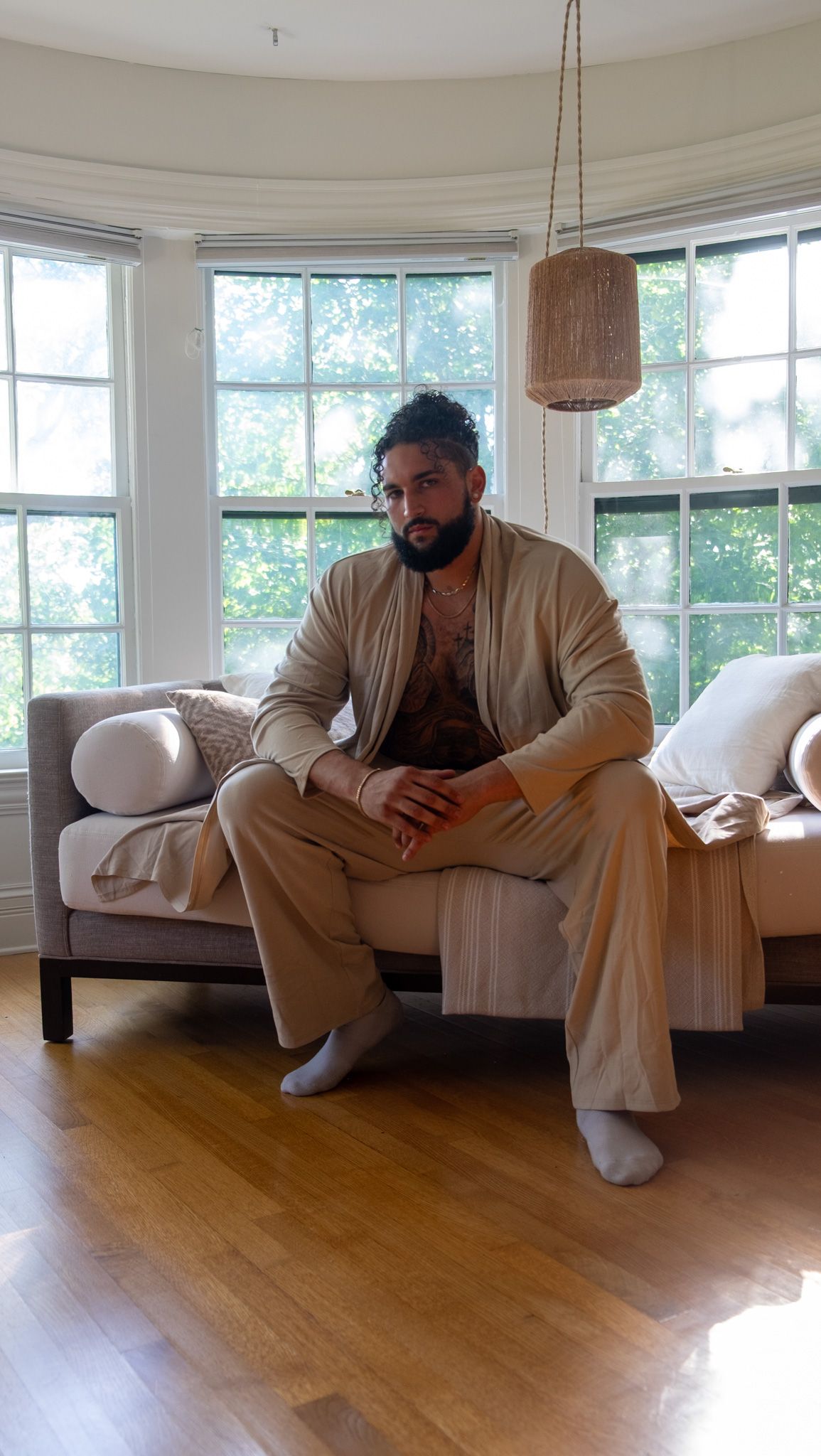 Bearded man in beige linen loungewear and socks sitting on a cushioned daybed in a bright bay-window room with hardwood floors.