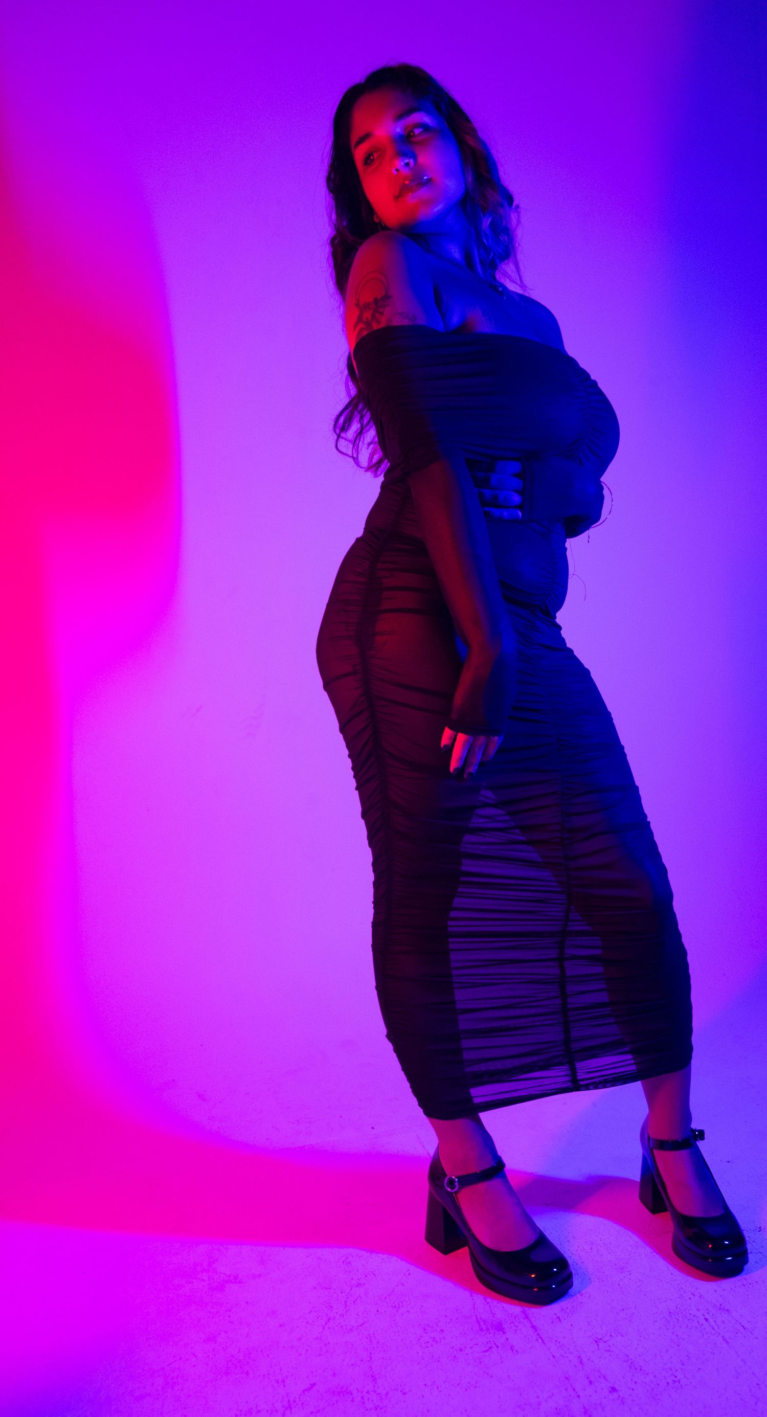 Woman in a black ruched sheer midi dress and platform Mary Janes posing in vivid pink and purple lighting in a studio.