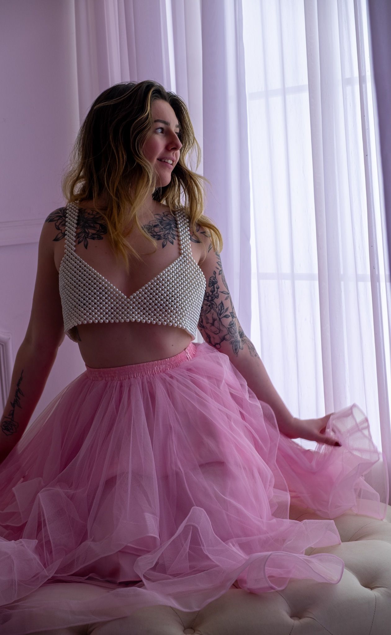 Smiling woman with tattoos in a pearl bralette and pink tulle skirt sits near sheer curtains in a soft lavender room.