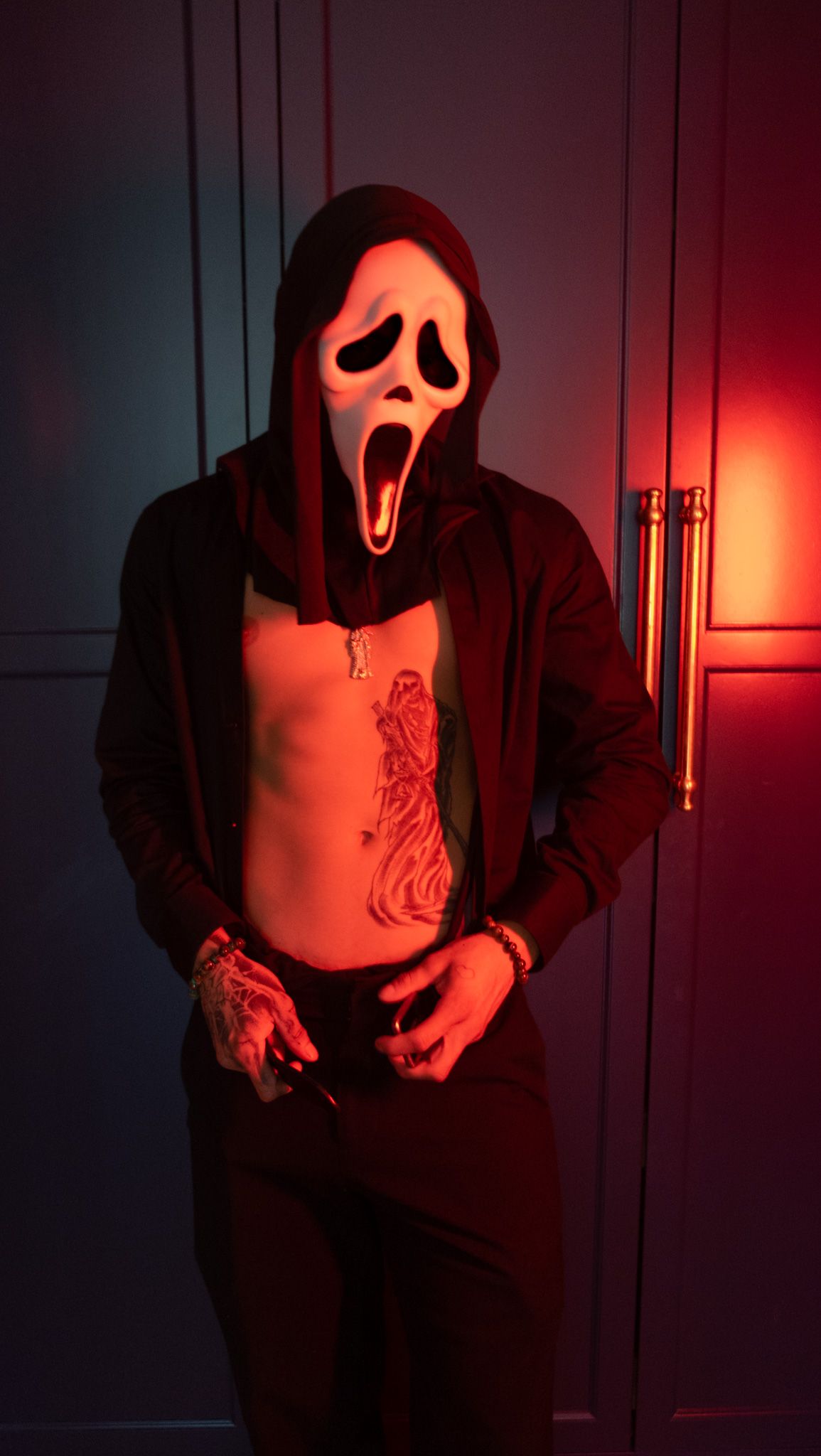 Person in a Ghostface mask and open dark shirt with a torso tattoo stands against green cabinets in red light.