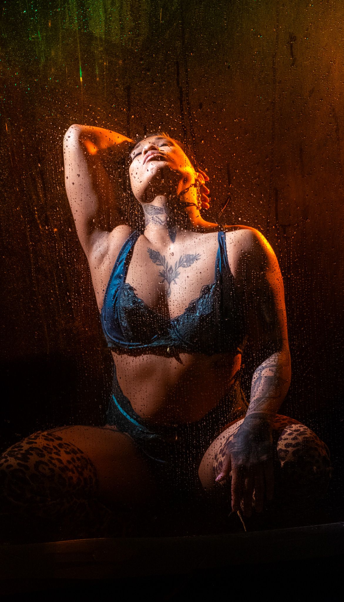 Tattooed woman in blue lace lingerie and leopard print stockings tilts her head back behind a wet, droplet-covered glass in warm light.