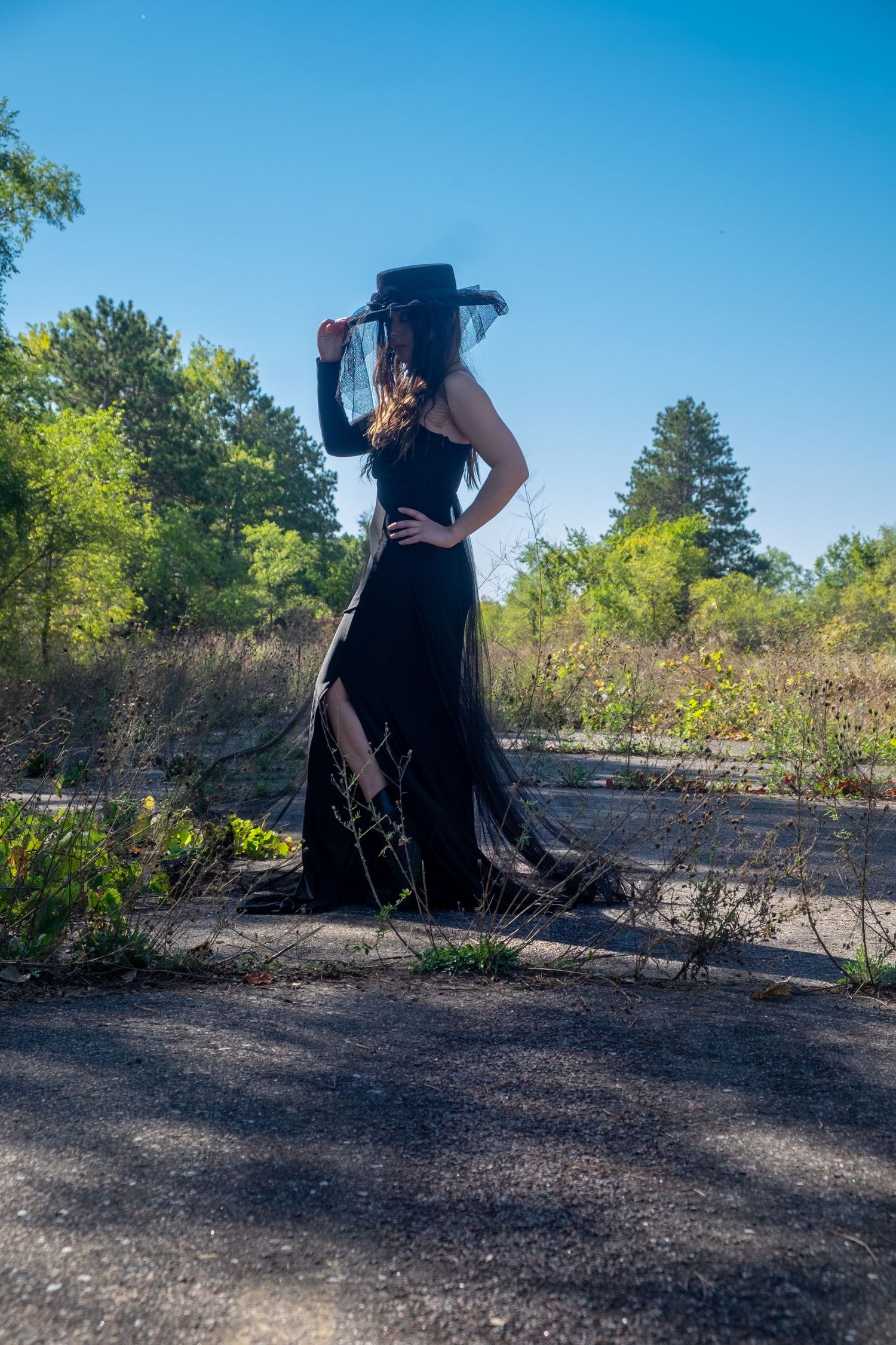 Woman in a long black dress and wide-brimmed hat with a veil poses on cracked pavement surrounded by trees under a blue sky.