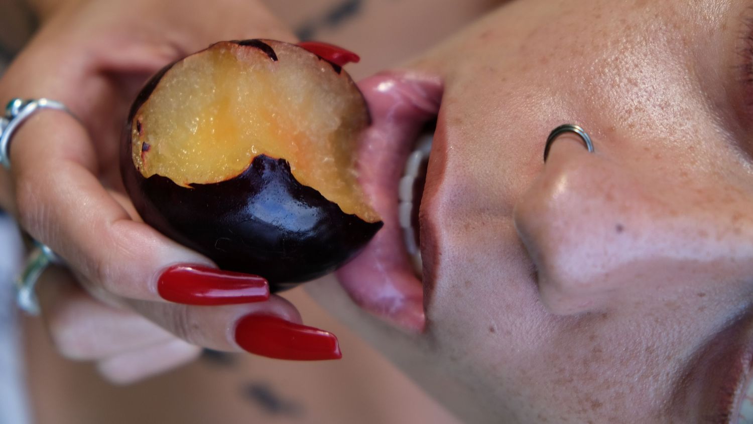 Close-up of a woman with red nails and a nose piercing biting into a juicy halved plum.