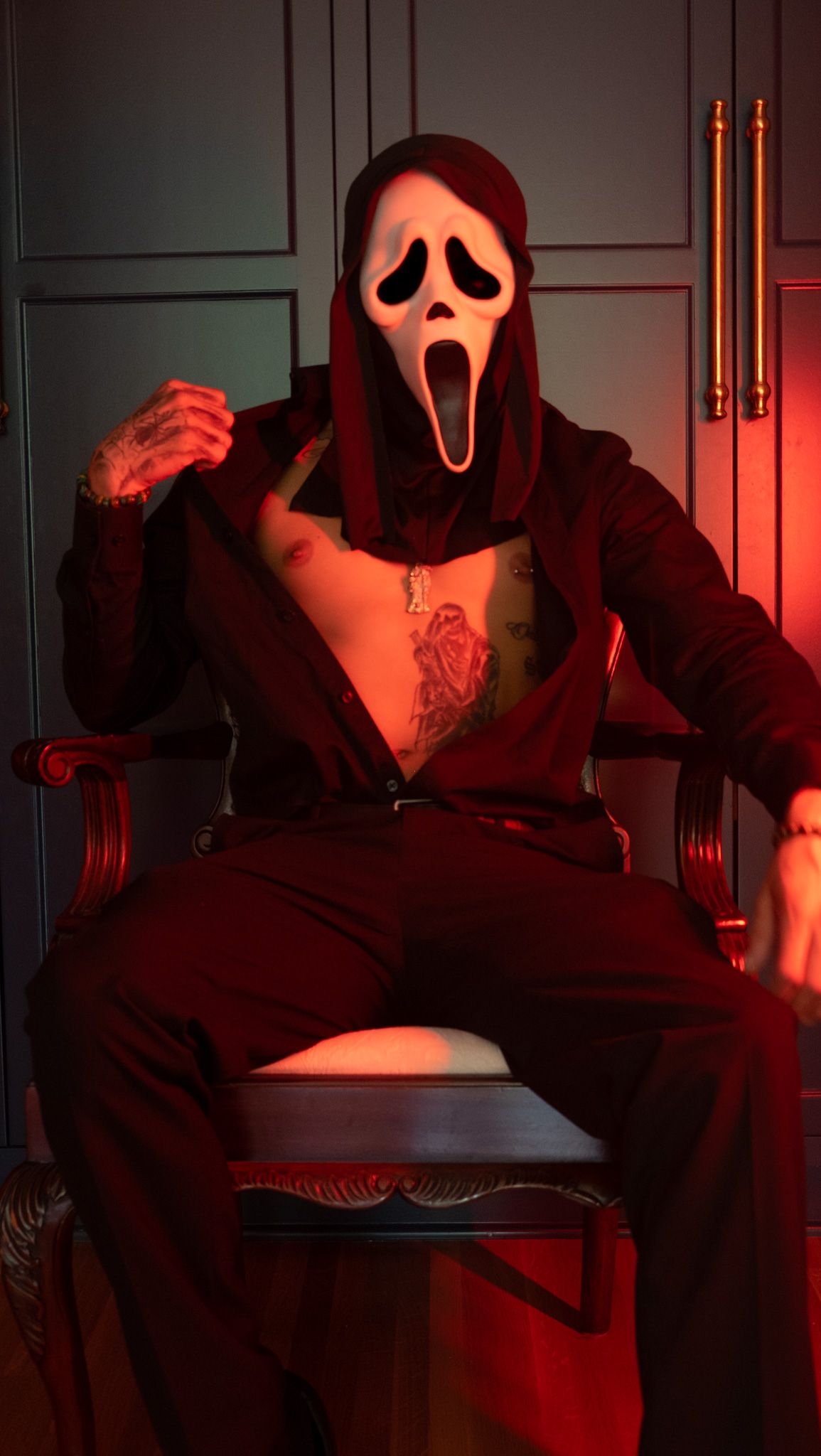 Person in a Ghostface mask and open dark shirt with chest tattoos sits in a wooden chair under moody red lighting.