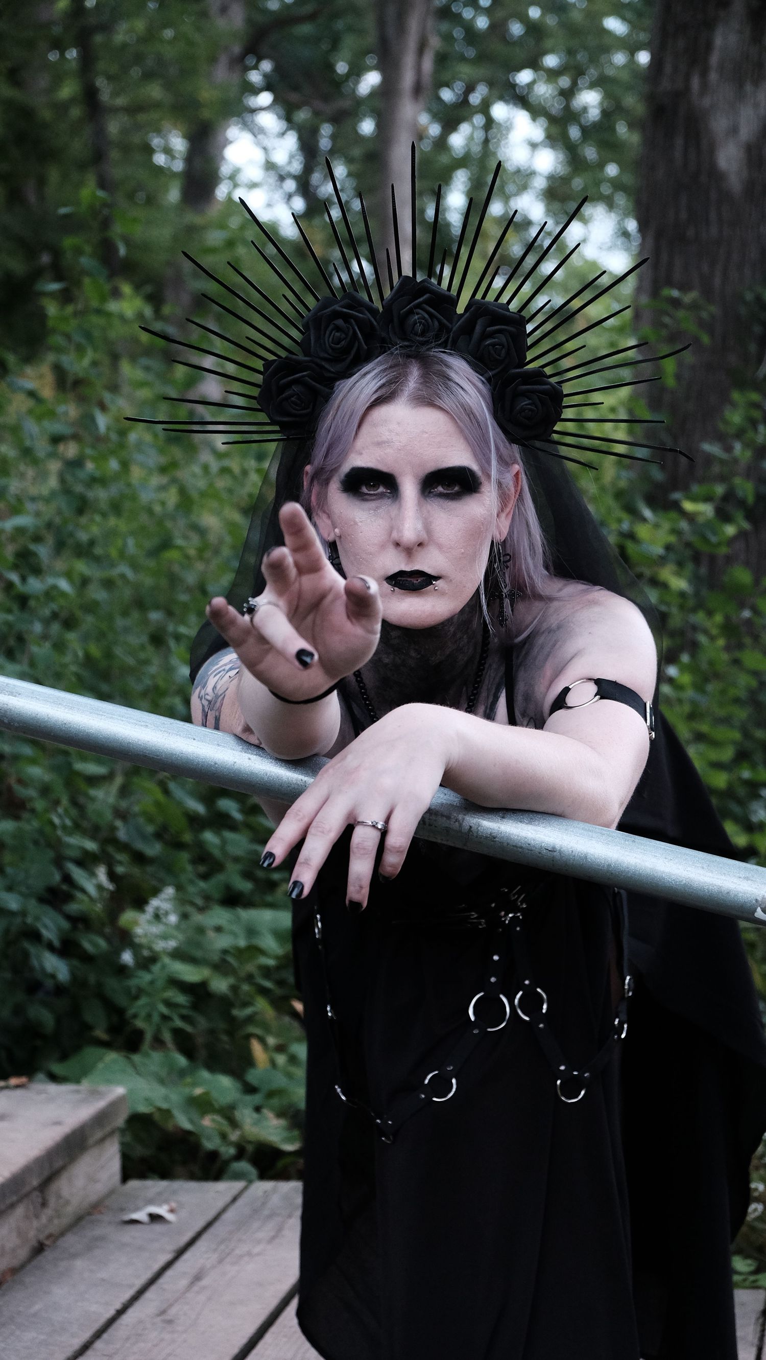 Gothic woman with a black rose spiked crown, dark makeup and black lipstick reaching toward the camera on a wooden deck in a forest.
