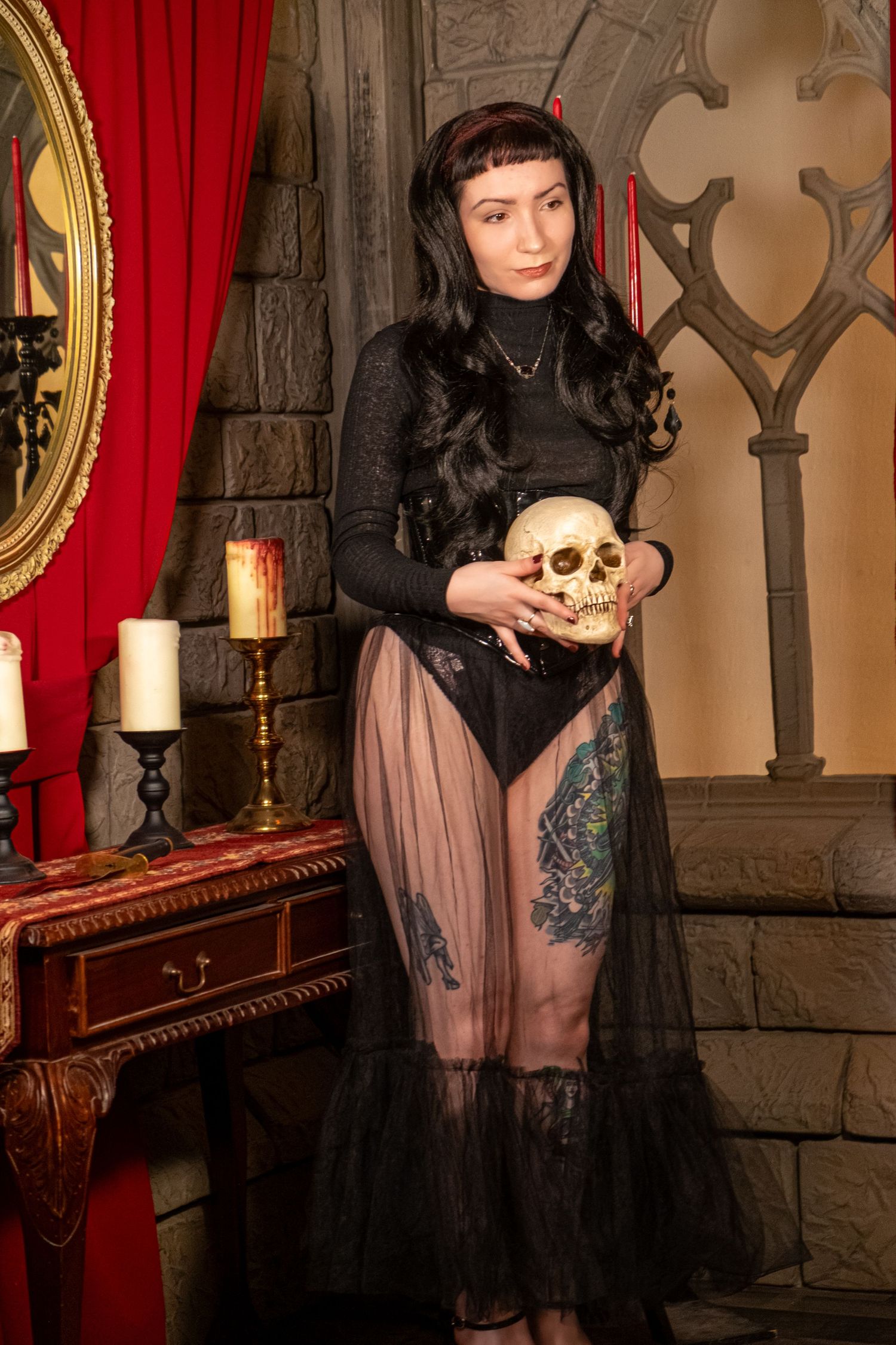 Woman with long black hair and leg tattoos holds a skull beside candles in a gothic room with stone walls.