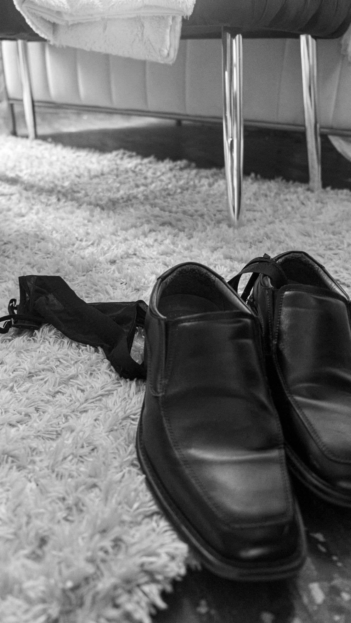 Black and white close-up of polished black dress shoes and a dark garment on a shaggy rug near a chrome-legged bench.