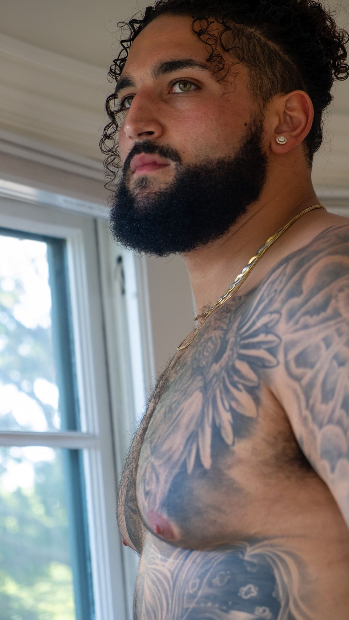 Close-up of a bearded man with curly hair, a gold chain and detailed chest and shoulder tattoos standing near a sunlit window.