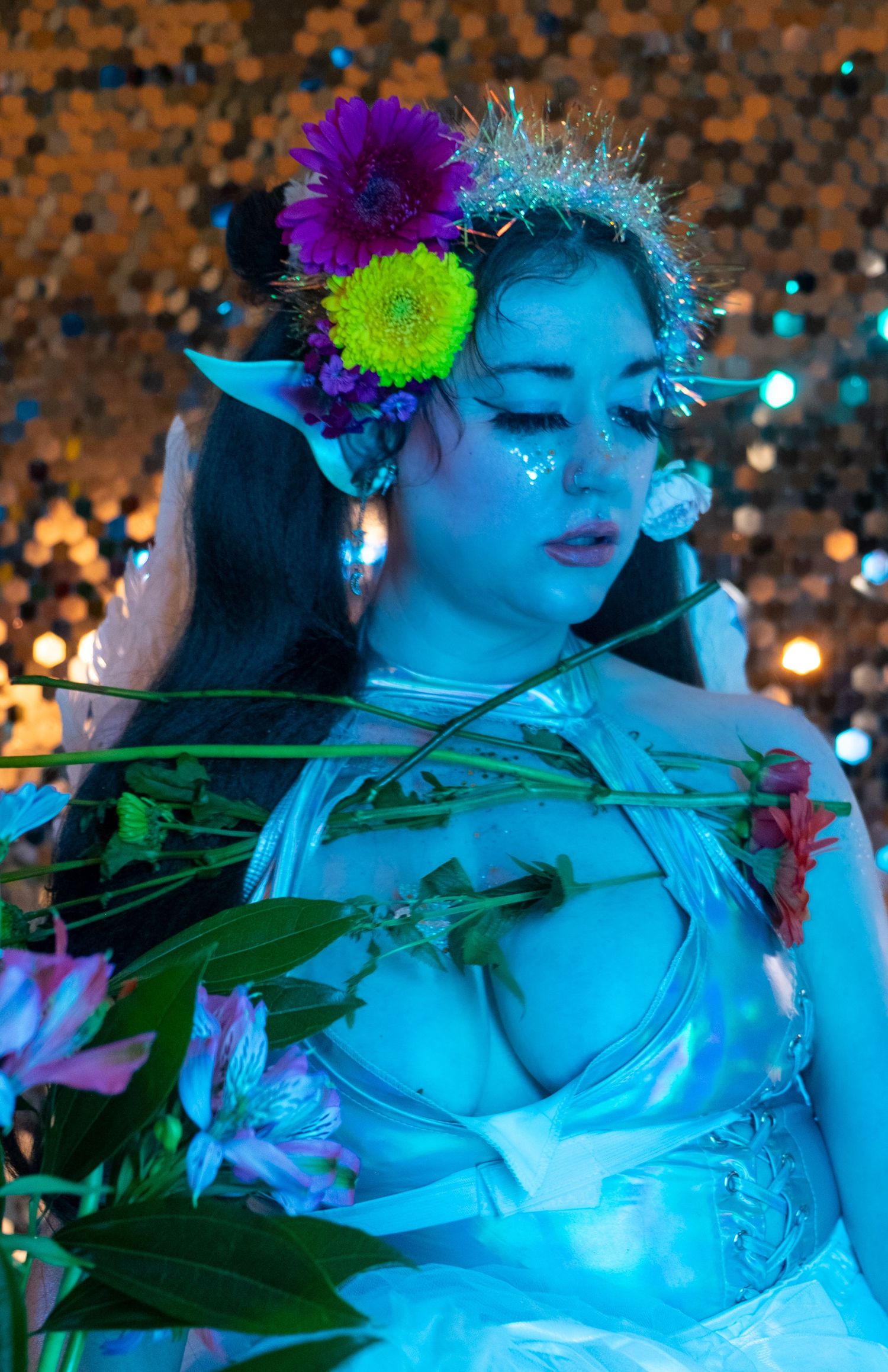 Close-up of a person in an elf costume with pointed ears, flower crown and glitter makeup holding flowers before sequins.