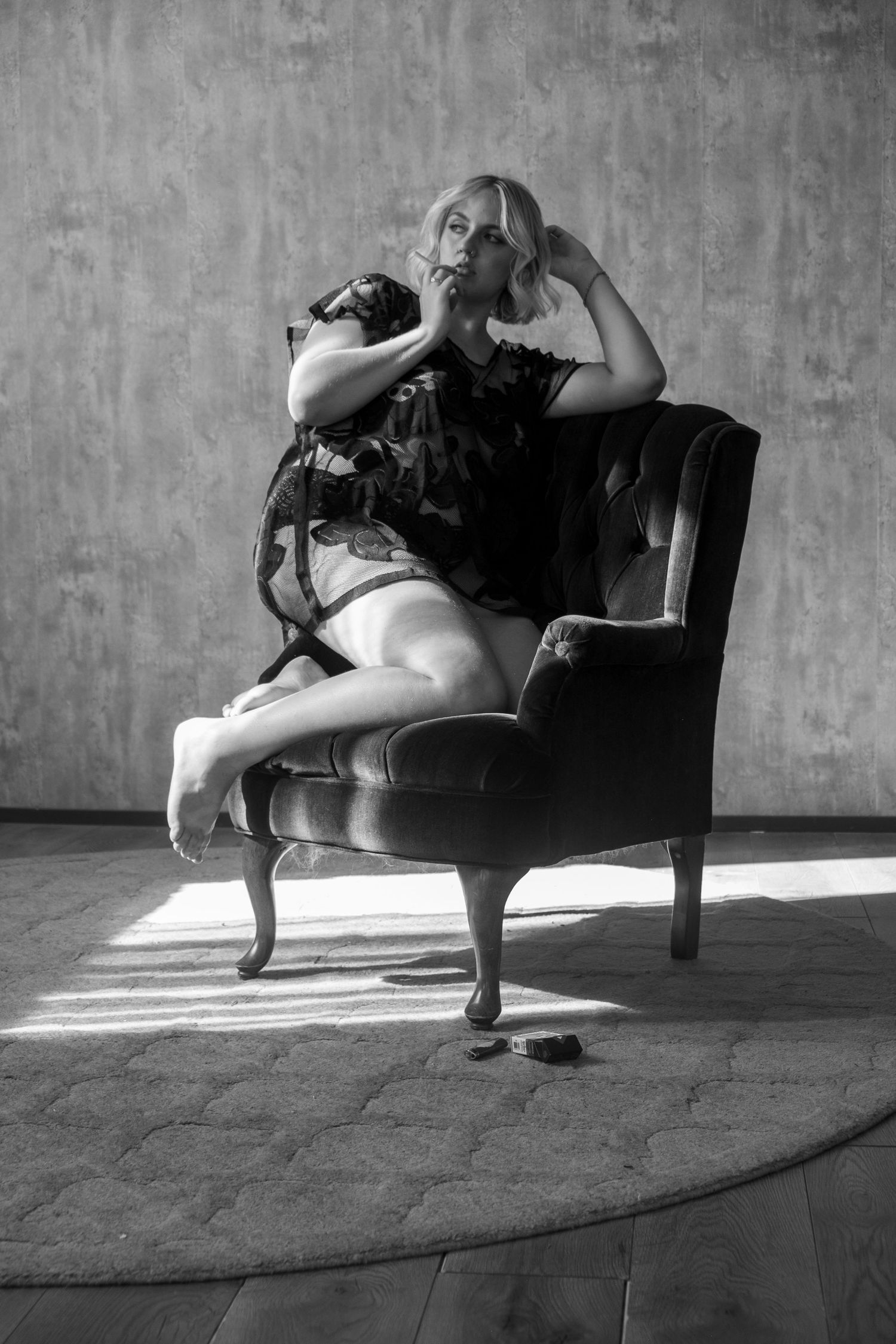 Black and white portrait of a woman in a sheer dark top seated sideways on a vintage armchair in soft window light.