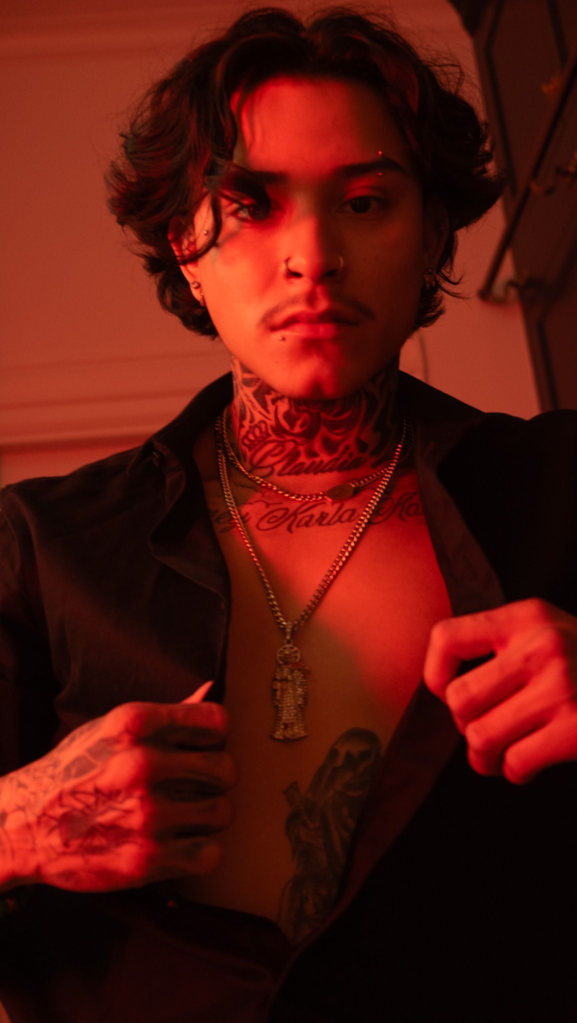 Tattooed man with neck tattoos, nose ring and gold chain necklaces opening his black shirt under dramatic red light.