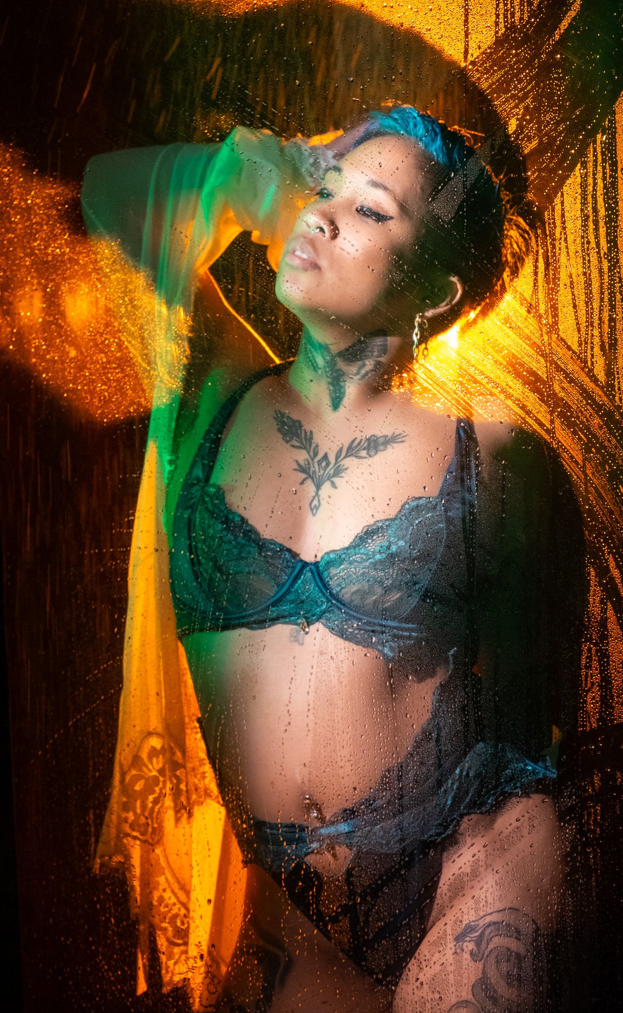Tattooed woman in teal lace lingerie poses behind a rain-streaked glass pane lit by vivid orange and green light.