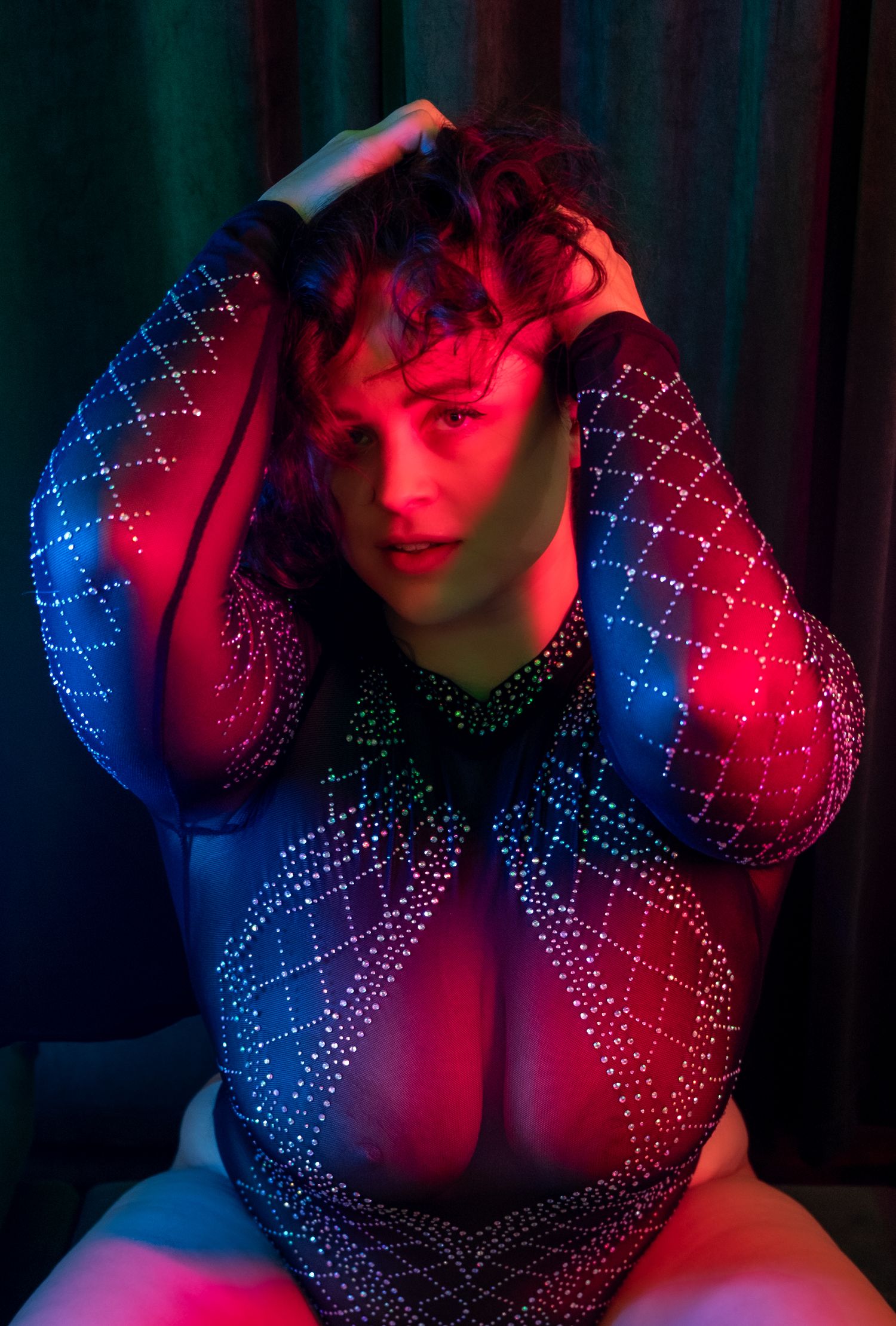 Person in a sparkly rhinestone mesh bodysuit poses with hands in curly hair under dramatic red and blue lighting.