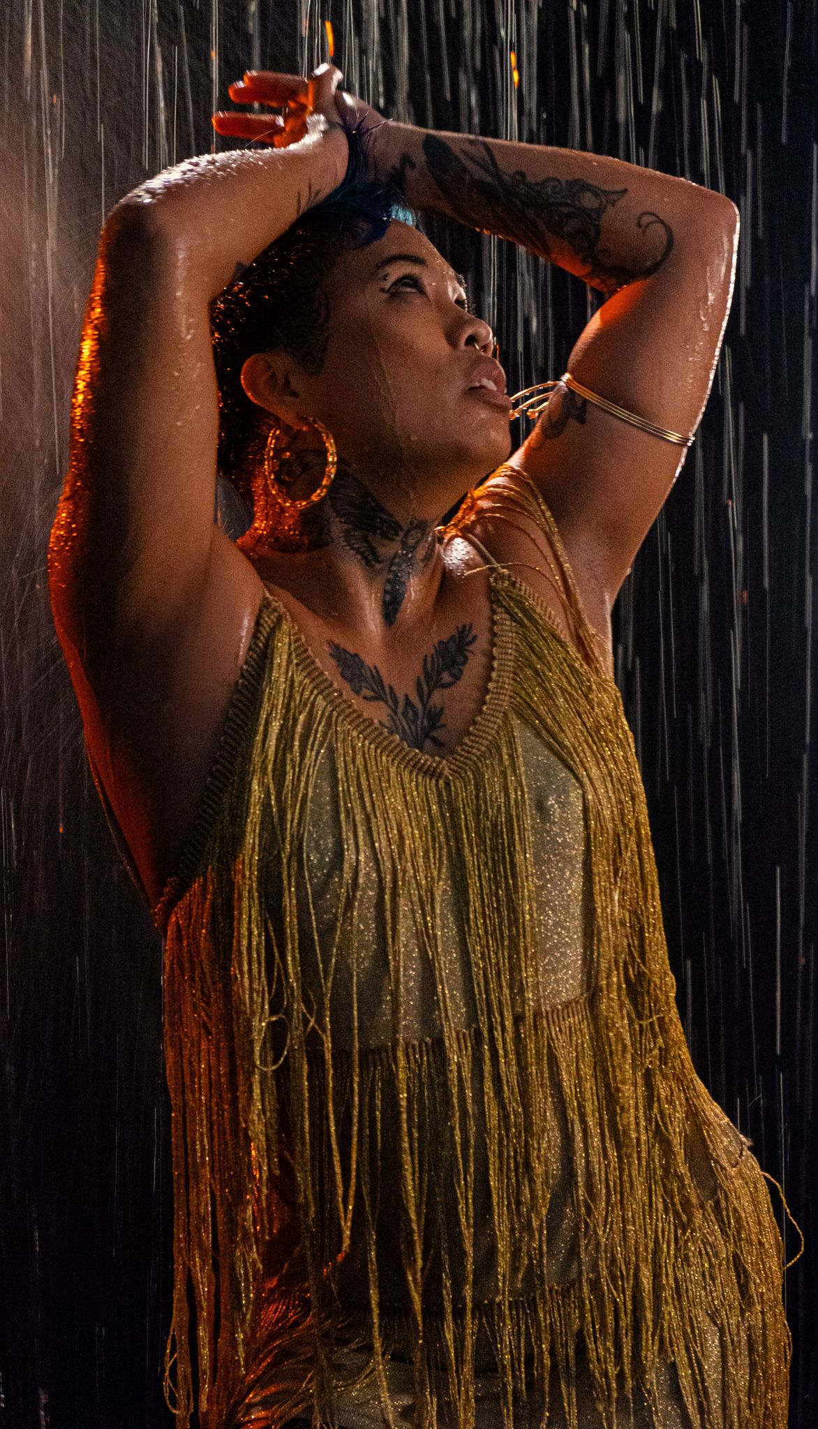 Tattooed woman in a gold fringe top and hoop earrings raises her arms in the rain under dramatic amber lighting.