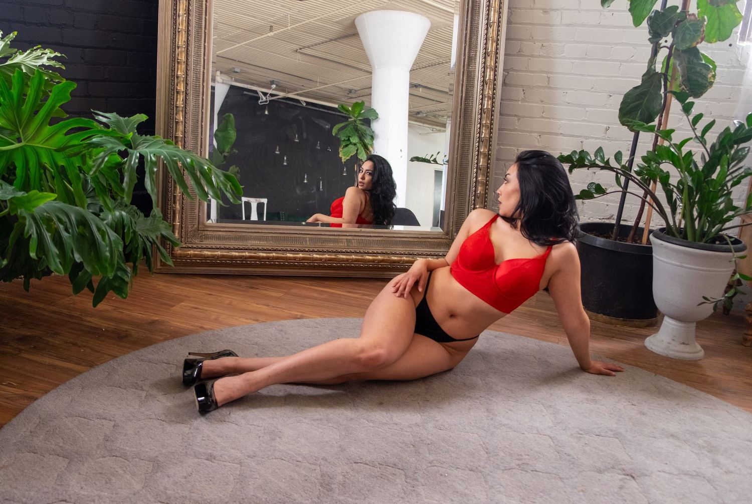 Woman in red top and black briefs with heels reclines on a gray rug beside a large gilded mirror and leafy potted plants.