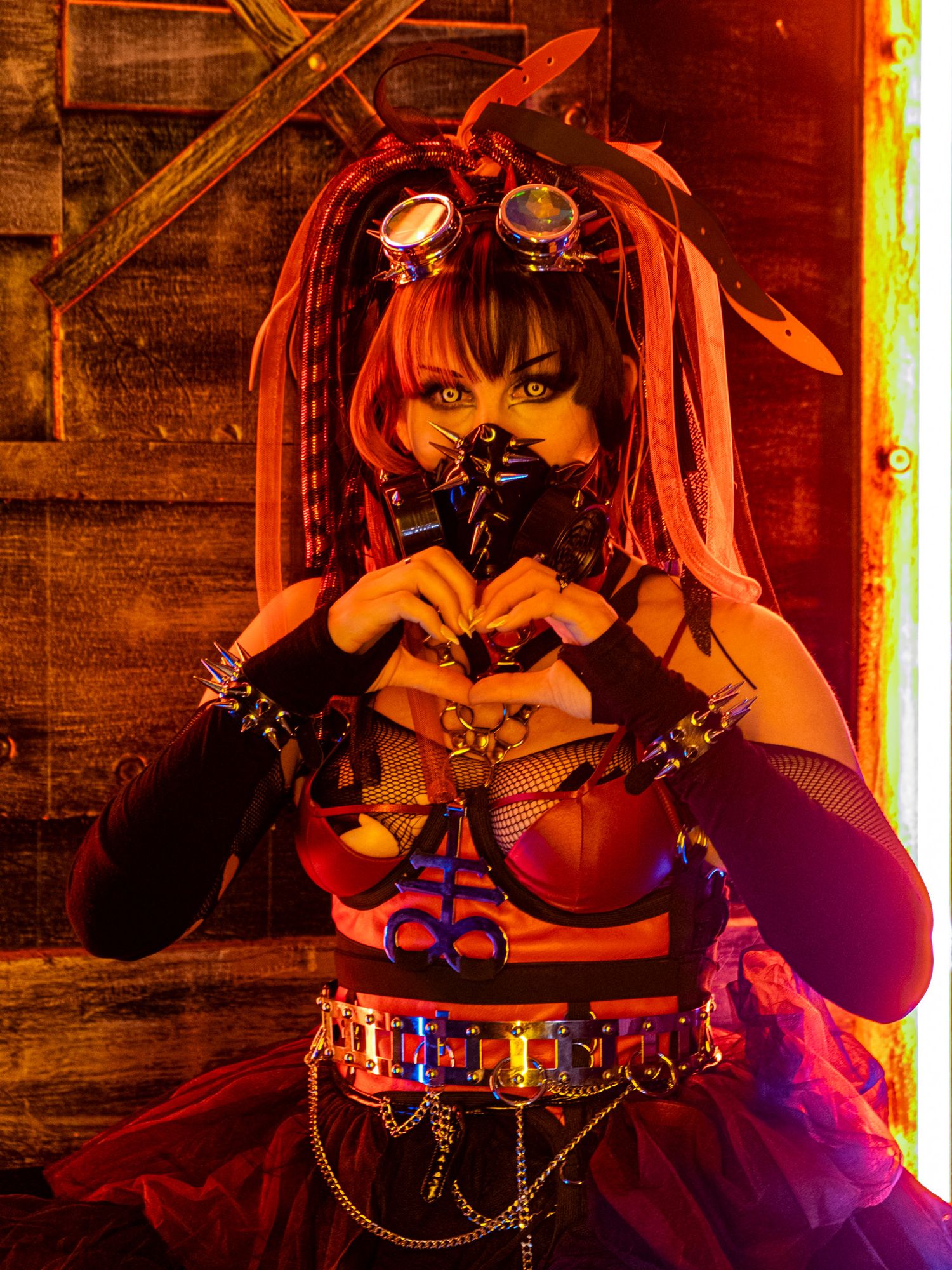 Cyberpunk-styled woman with goggles, red and black dreadlock hair, face mask and spiked gloves making a heart shape with her hands.