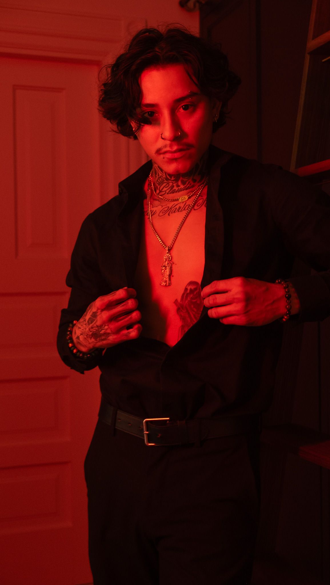 Tattooed man with layered gold necklaces unbuttons a black shirt in a red-lit room beside a white paneled door.