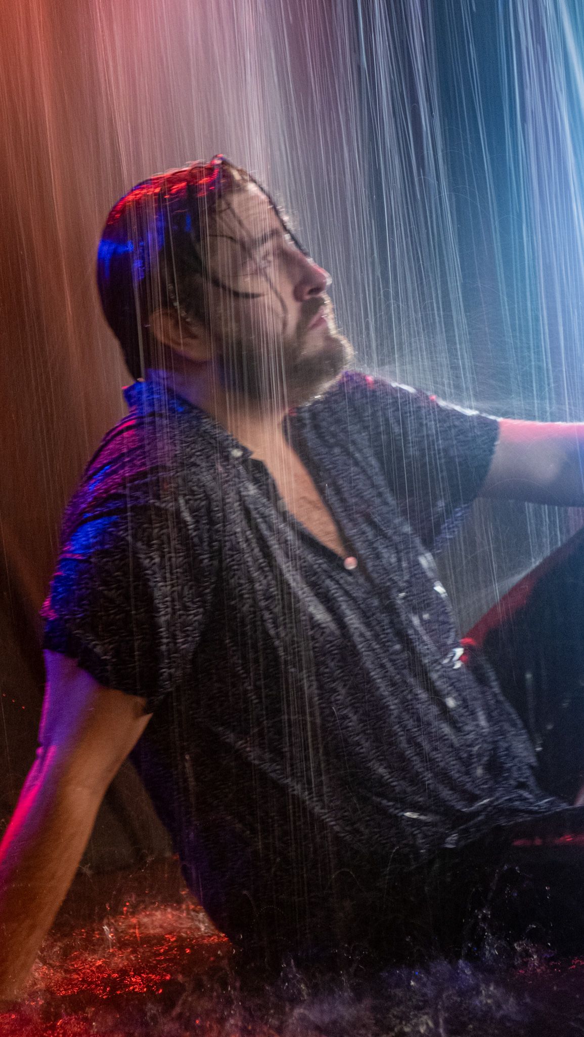 Drenched man with a beard gazes upward as rain pours down around him in dramatic blue and red studio lighting.