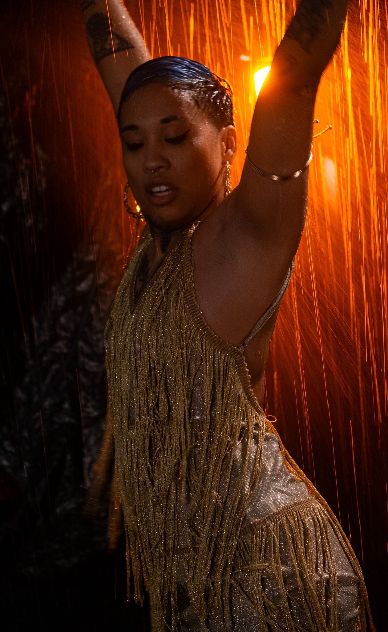 Woman with tattooed arms dances under falling water in a gold fringe top, lit by warm orange light.