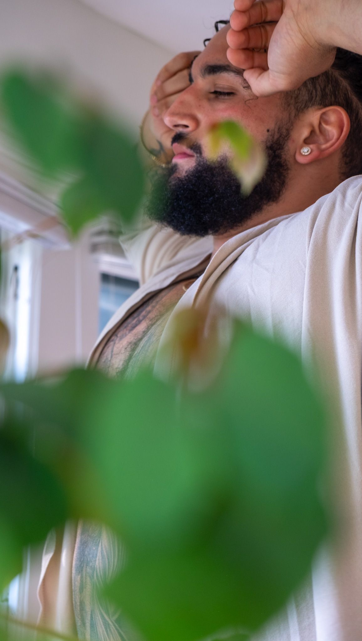 Close-up of a bearded man with earring resting his arm over his head, framed by blurred green leaves in soft light.