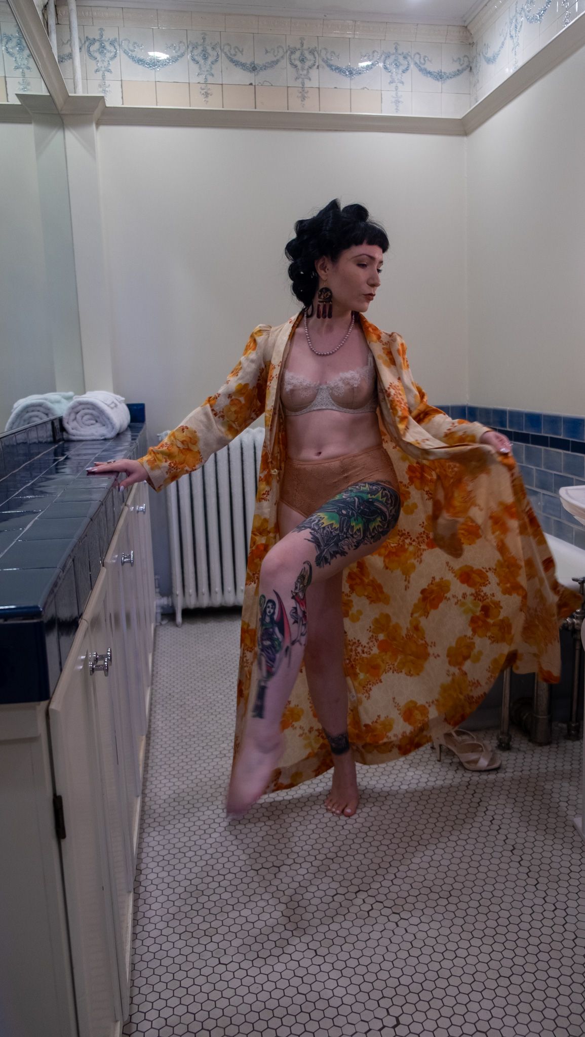 Tattooed woman with an updo in a floral robe and lingerie posing in a vintage tiled bathroom with blue and white tile.