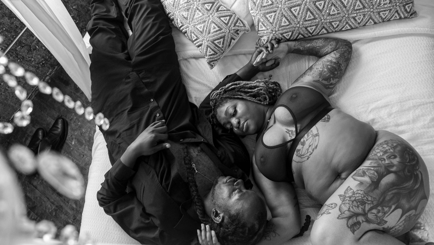 Black and white overhead photo of two people embracing as they lie together on a bed with patterned pillows.