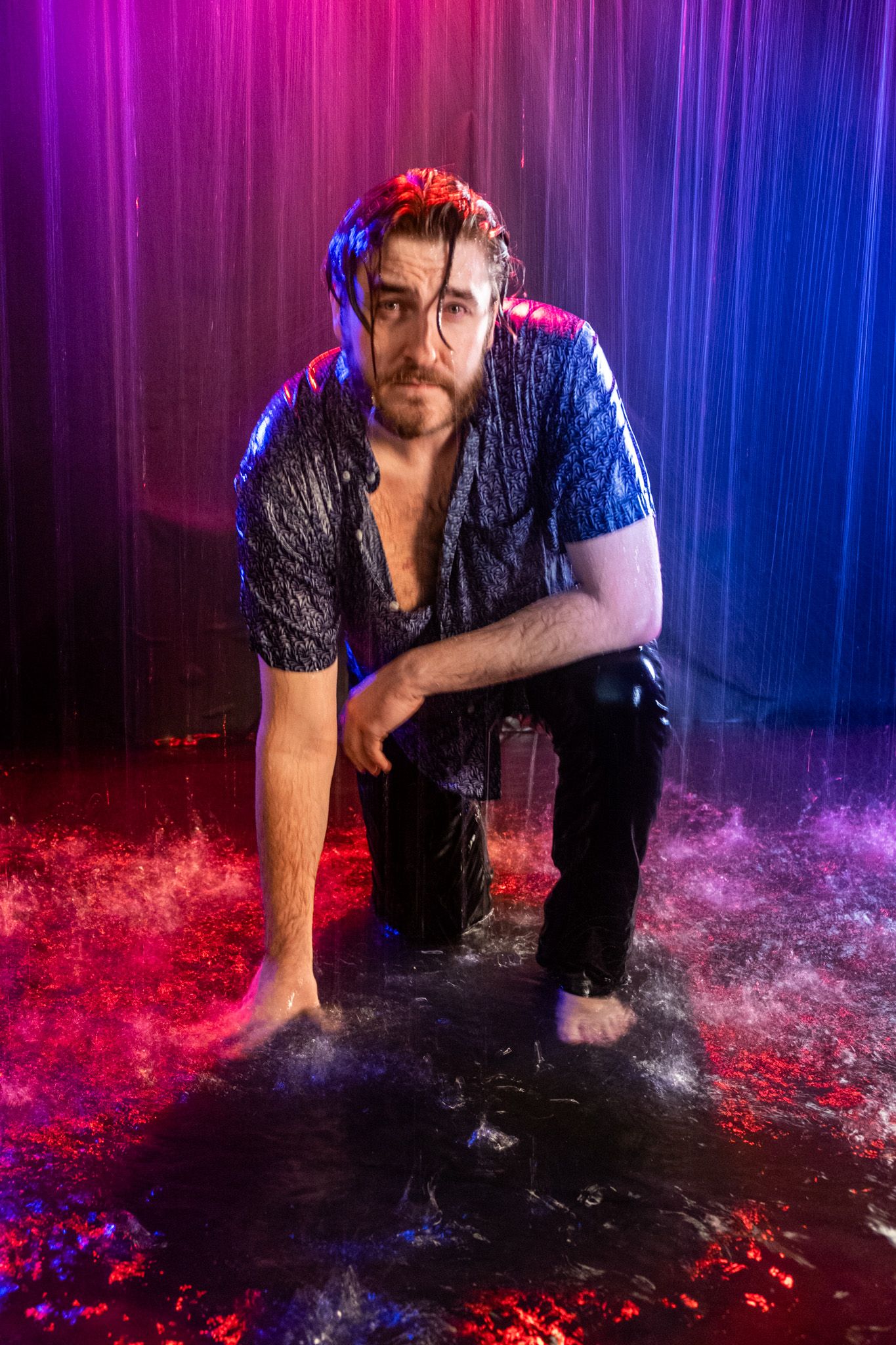 Wet bearded man in a dark shirt crouches barefoot in falling water under vivid pink and blue studio lighting.