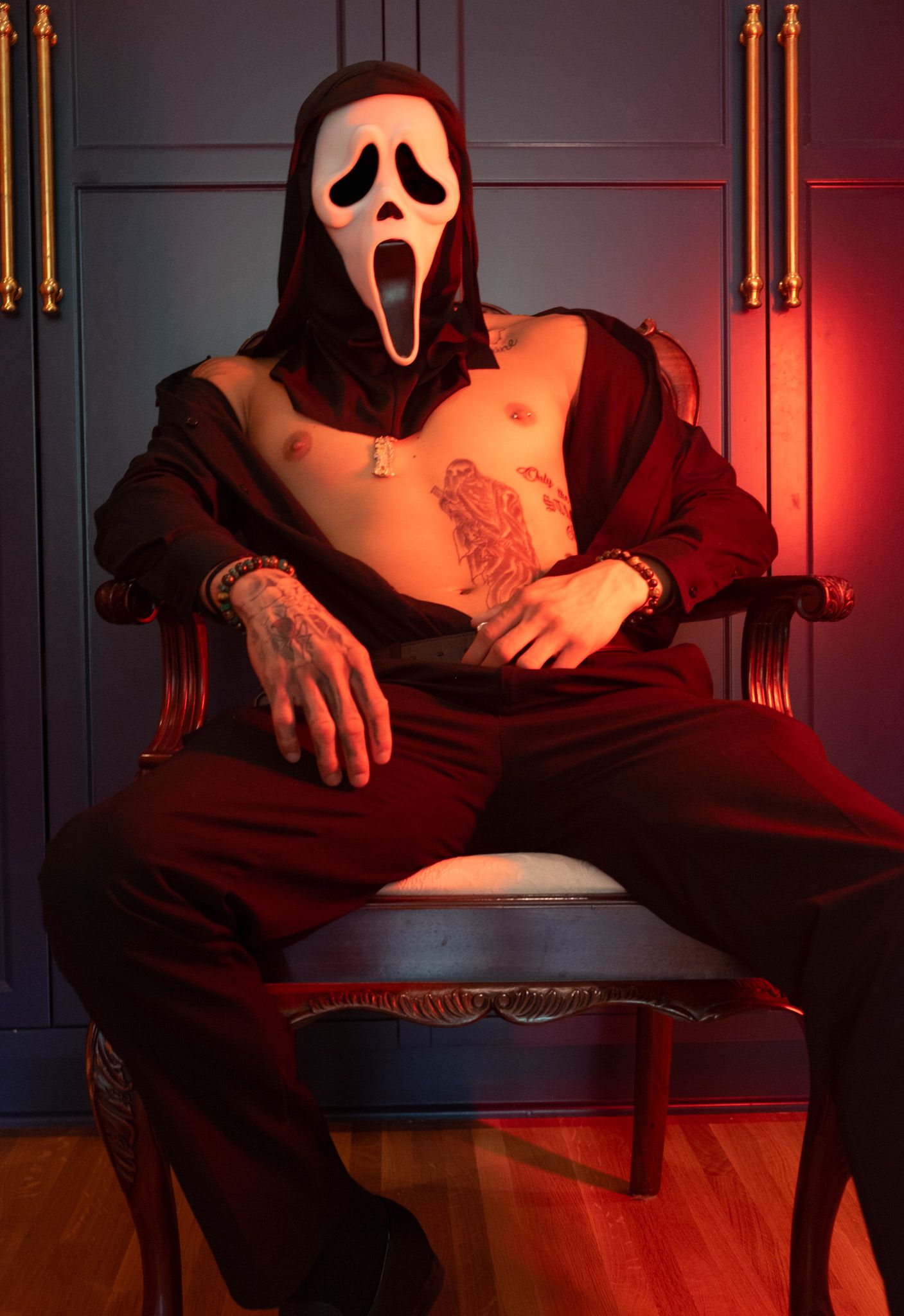 Tattooed man in a white Ghostface mask and open dark red shirt sits in a wooden chair against dark blue cabinets in red light.