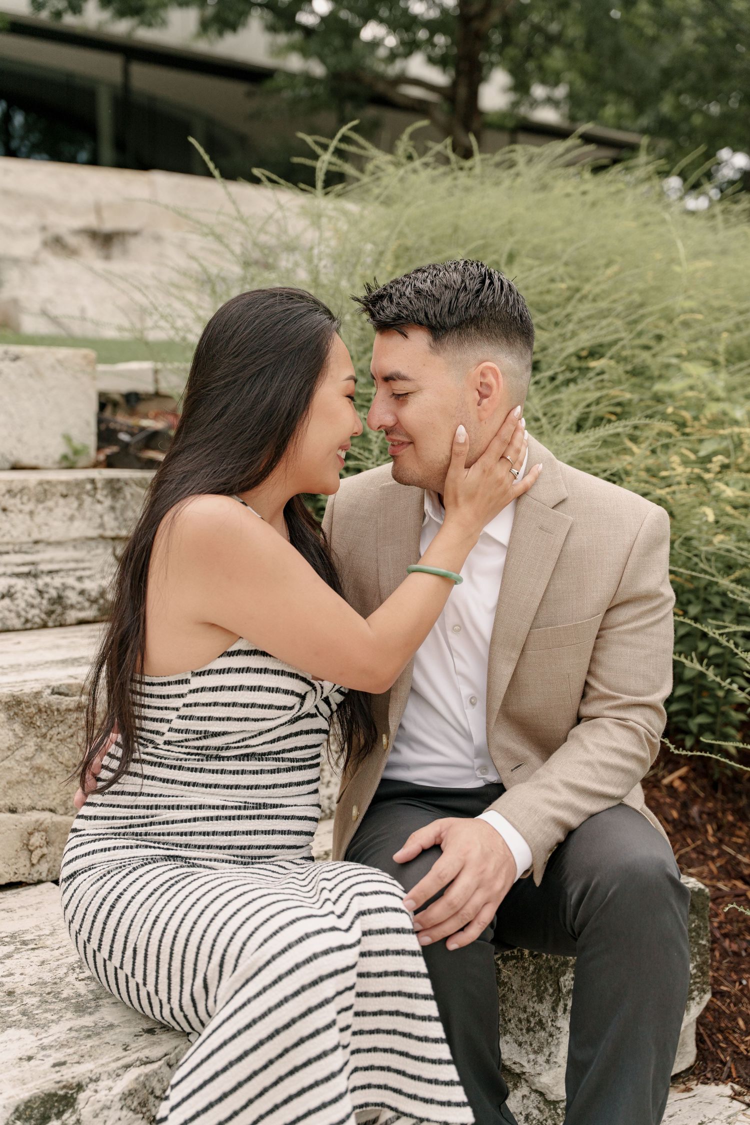 An engaged couple sit on stone steps touching foreheads and smiling, with the woman's hand gently on his face.