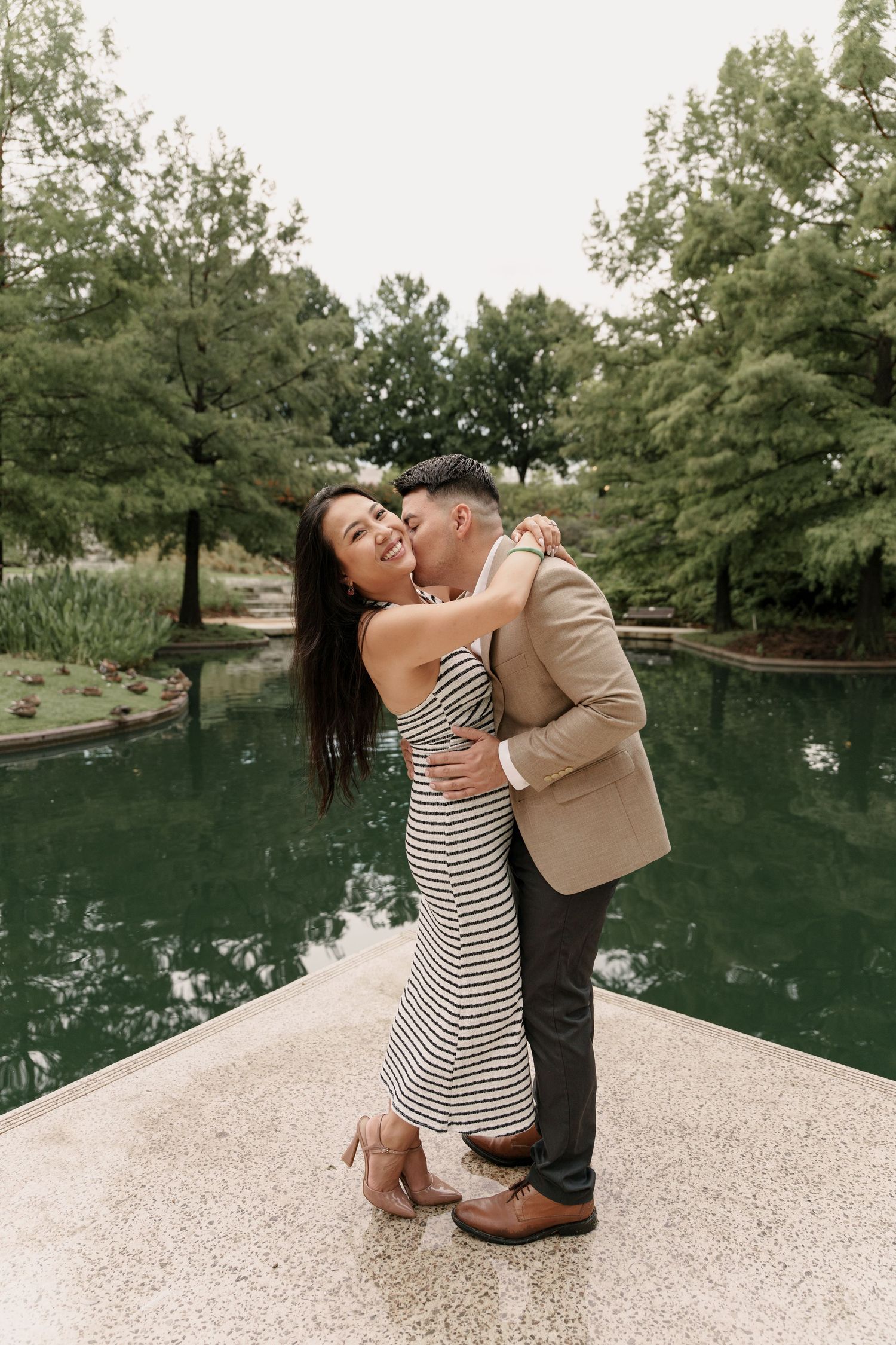 A woman in a striped dress laughs as her fiancé kisses her cheek on a stone path beside a green pond.