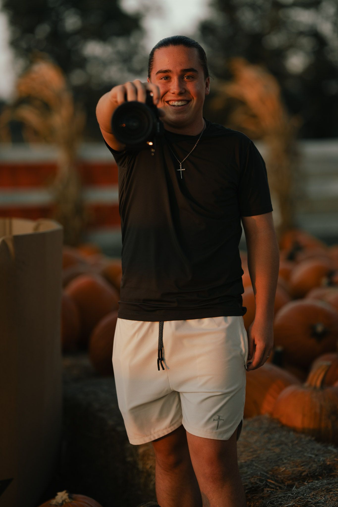 Smiling photographer in a black shirt and white shorts points his camera at the viewer while standing among pumpkins at sunset.