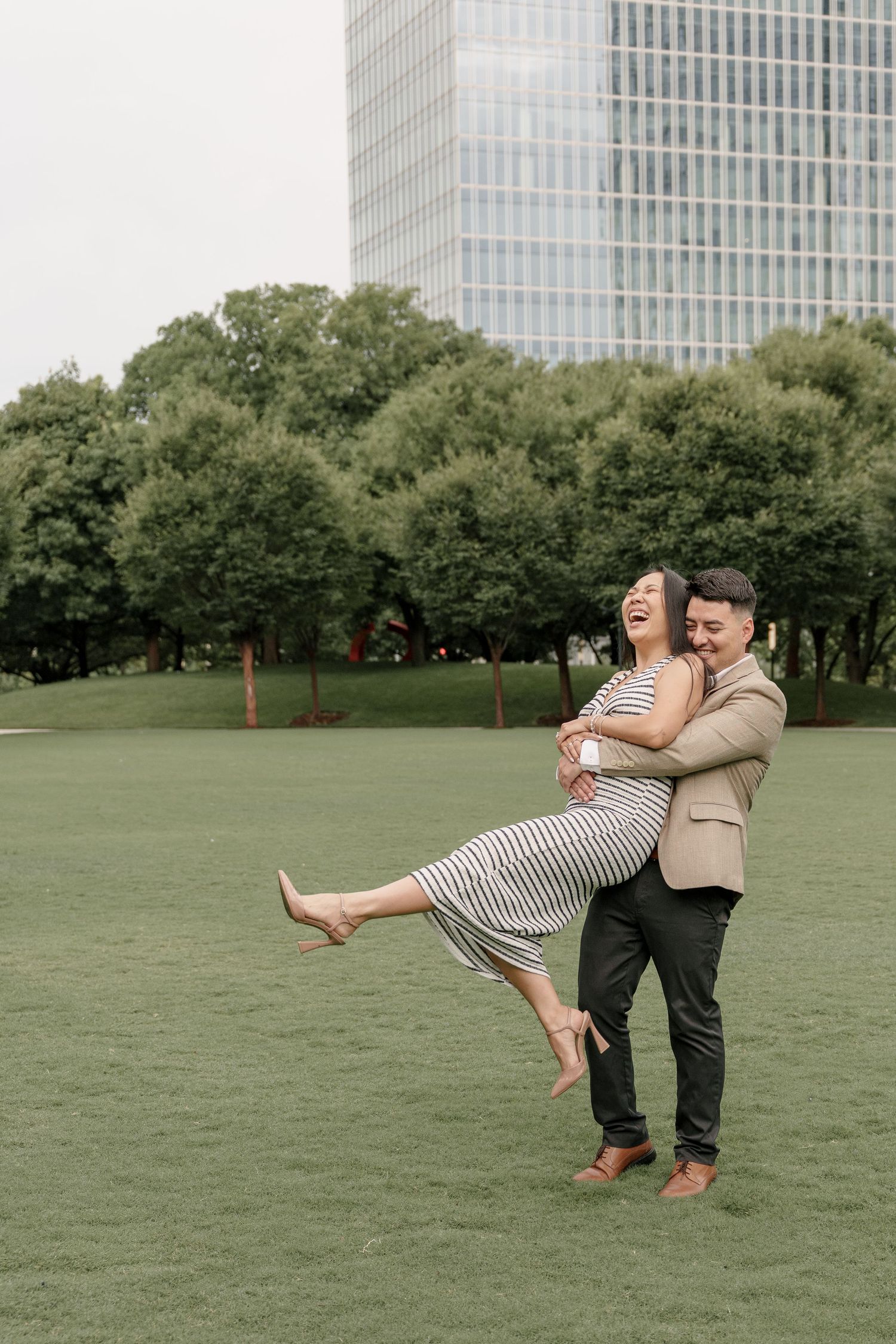 Man in a tan blazer lifts his laughing fiancée in a striped dress off the ground on a grassy lawn near tall city buildings.