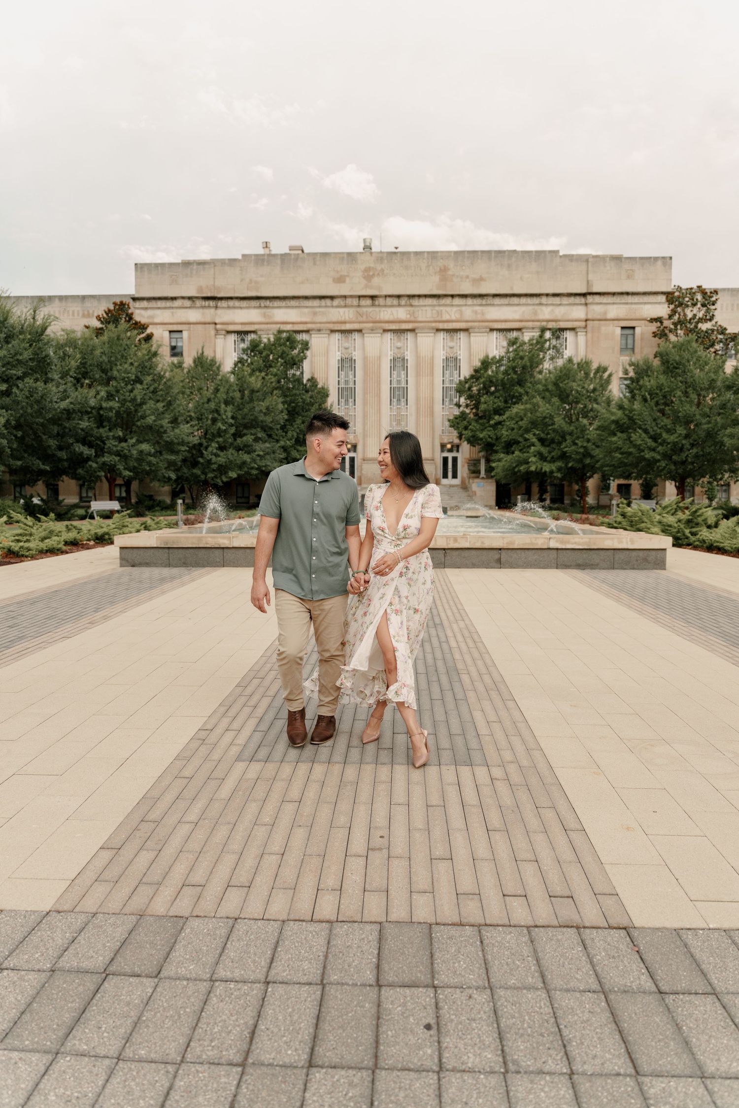 Engaged couple holding hands and walking across a brick plaza with a fountain and stately building behind them.