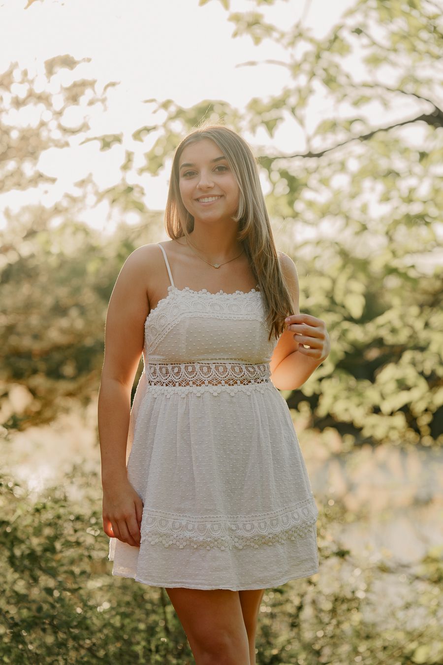 Smiling young woman in a white lace-trim sundress poses among green leafy trees for a natural light senior portrait.