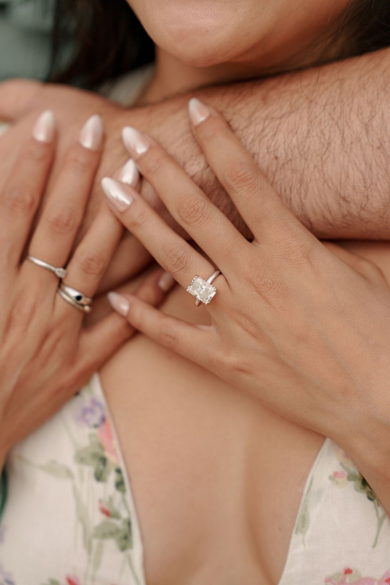 Close-up of a woman's hand showing a radiant-cut diamond engagement ring as a man's arm wraps around her.