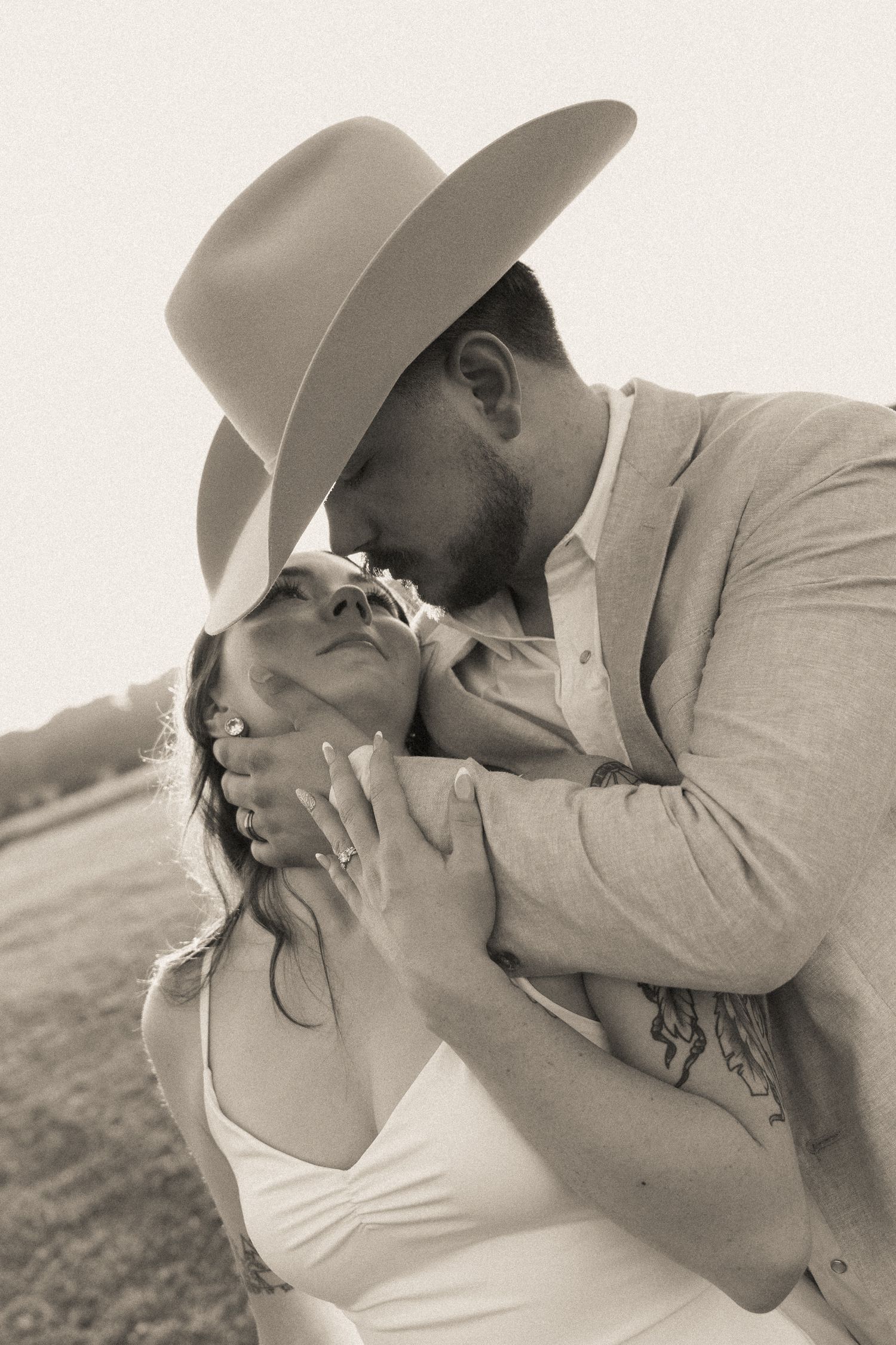 Black and white portrait of a groom in a cowboy hat kissing his fiancée's forehead as she looks up at him.