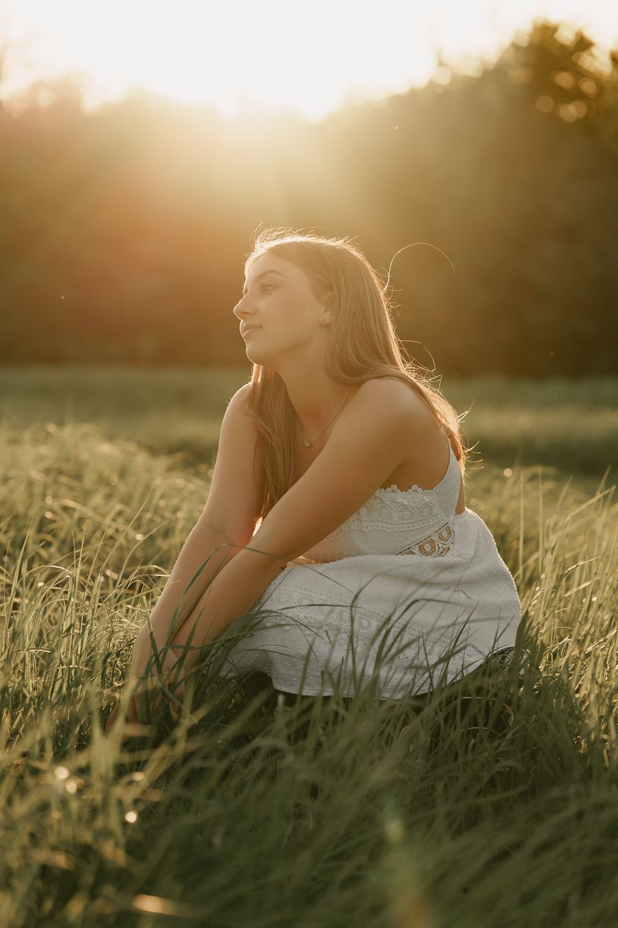 Woman in a white dress kneels in a sunlit field, gazing off to the side as the golden sun sets behind the trees.