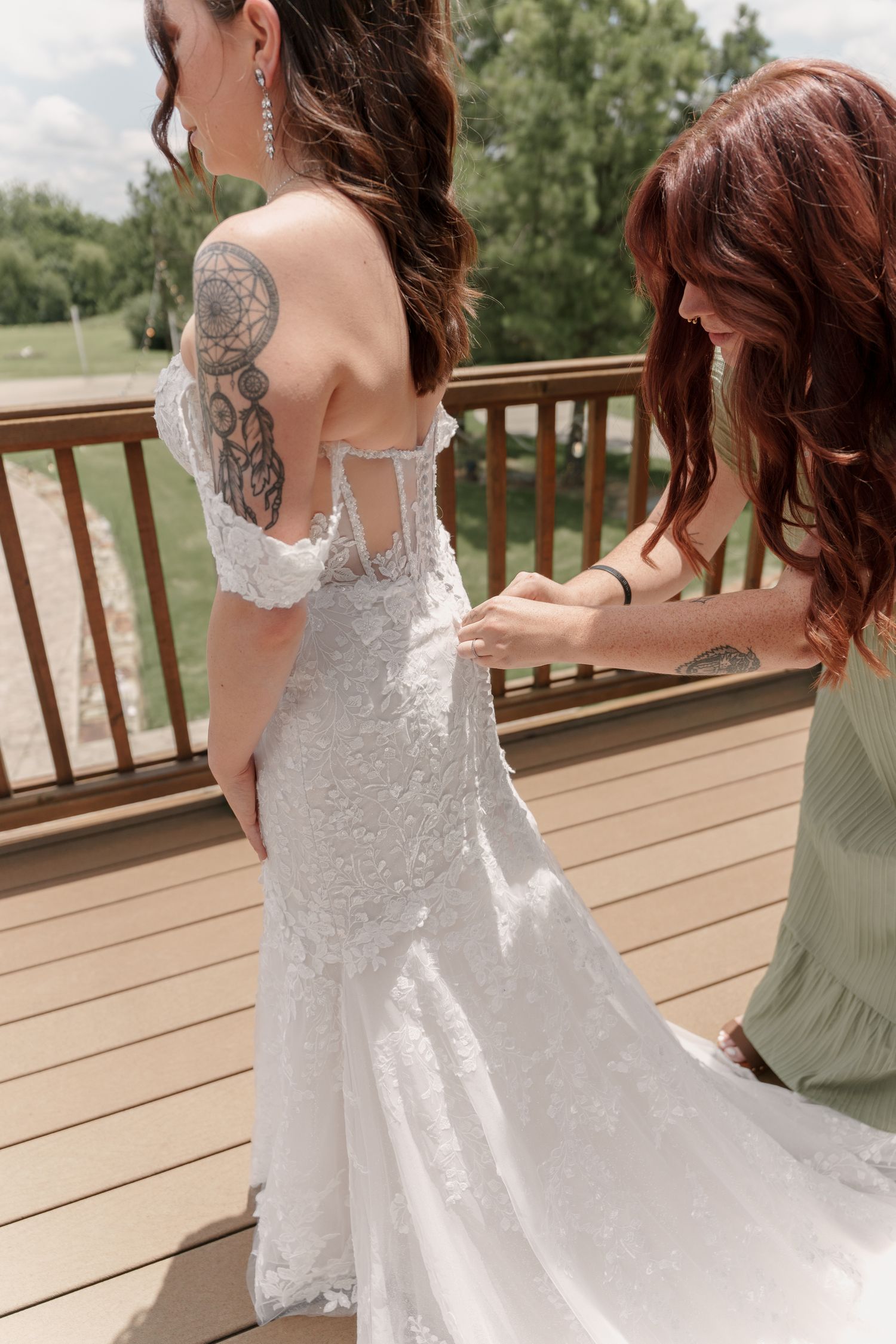 Bridesmaid in a sage green dress buttons the back of a bride's lace wedding gown on a wooden deck overlooking the countryside.