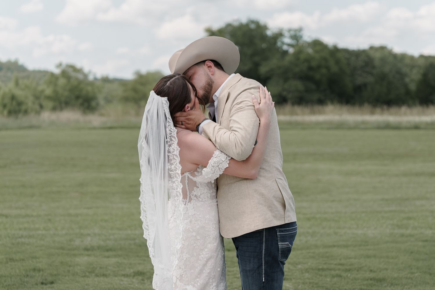 Bride in a lace gown and veil kisses her groom in a cowboy hat in a wide open grassy field.