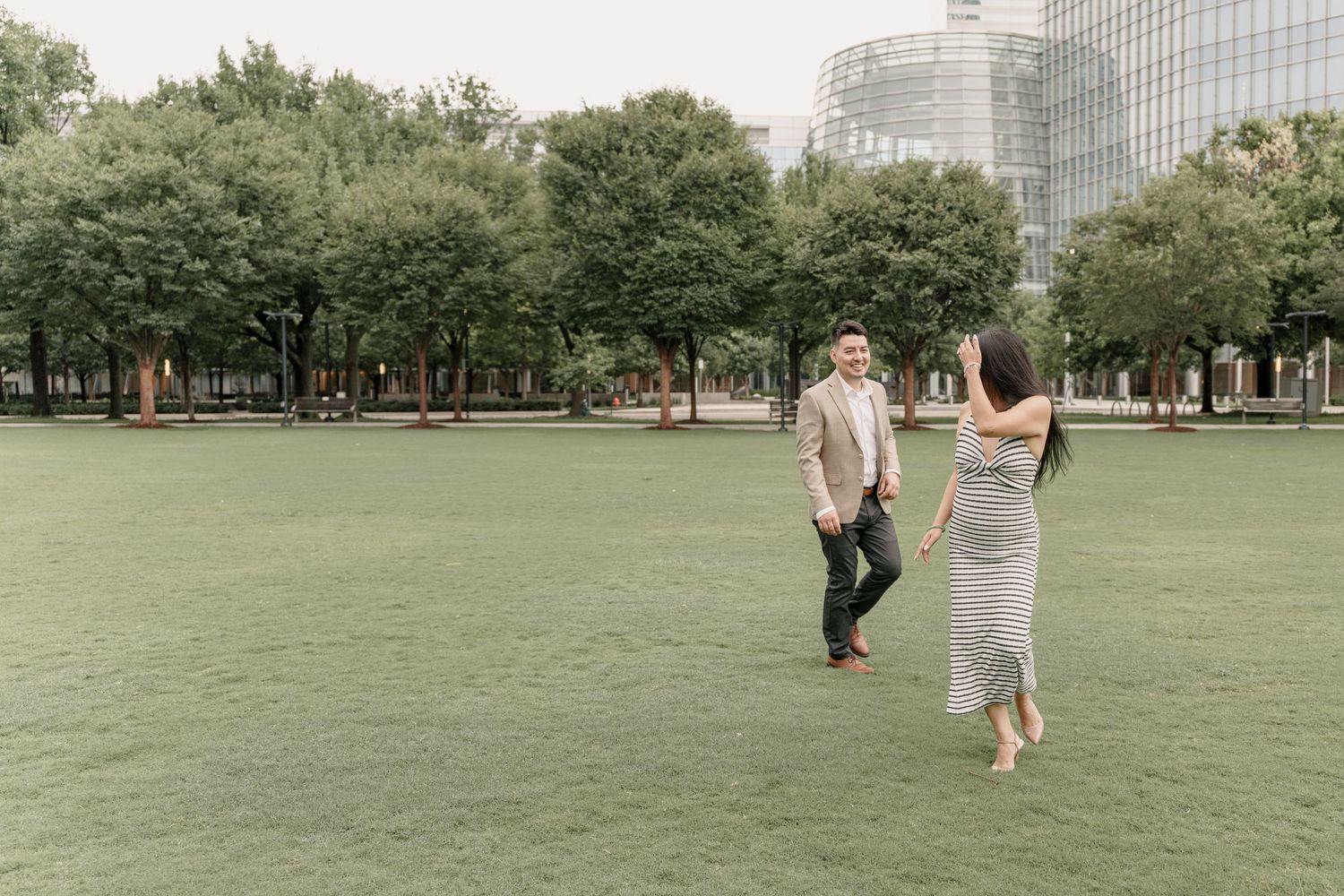 Smiling couple in a green park lawn with trees and a glass skyscraper behind them during an engagement photo session.