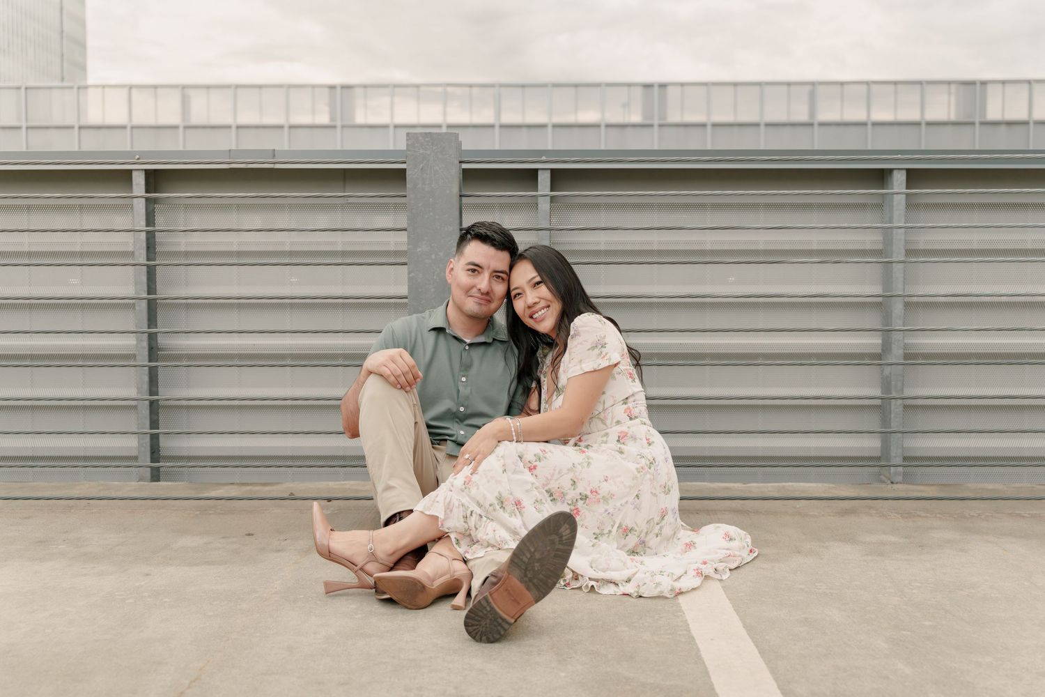 Couple sits close together on a rooftop parking deck, smiling and leaning in for a relaxed engagement portrait.