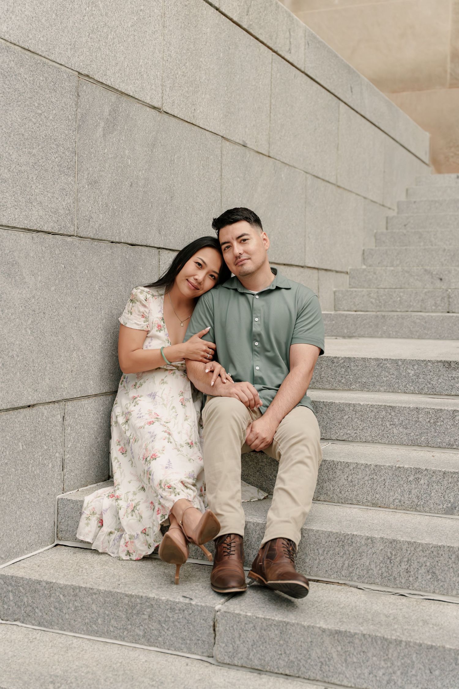 Engaged couple sits snuggled together on wide gray granite steps, she resting her head on his shoulder.