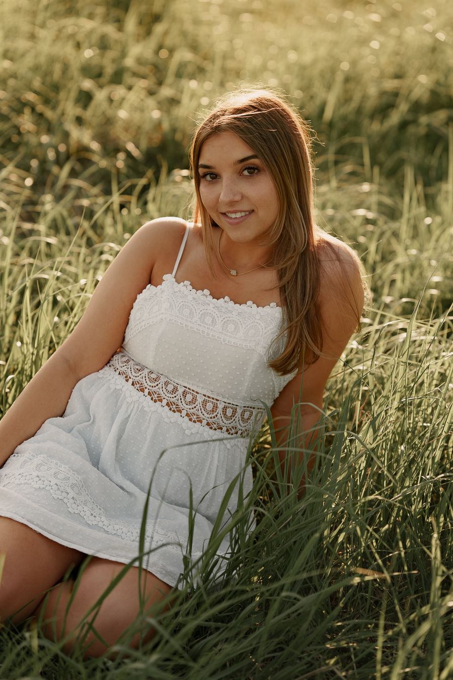 Young woman in a white sundress sits smiling in tall green grass during a warm golden hour outdoor senior portrait.