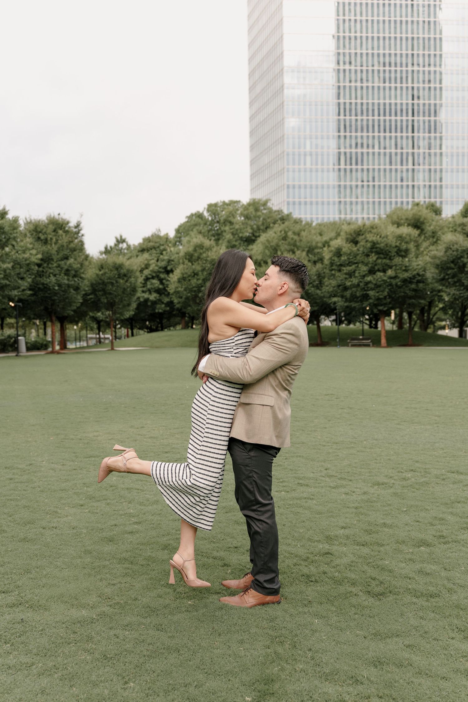 Couple kisses on a park lawn as the man lifts his fiancée, with trees and a glass high-rise in the background.