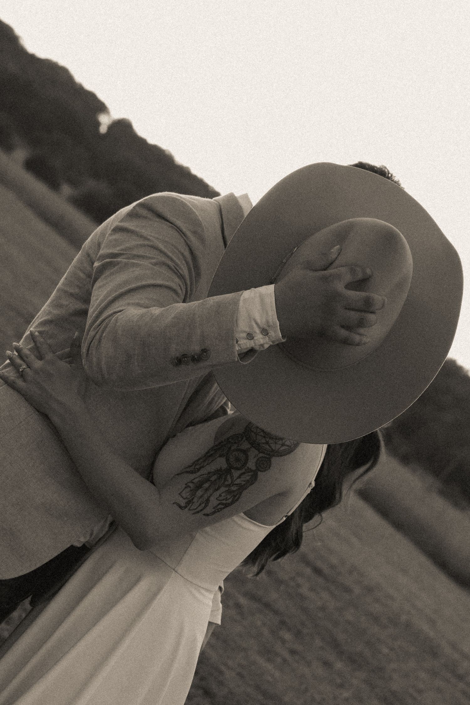 Black and white photo of a couple embracing as the man tilts a cowboy hat over their faces, with the woman's arm tattoo visible.
