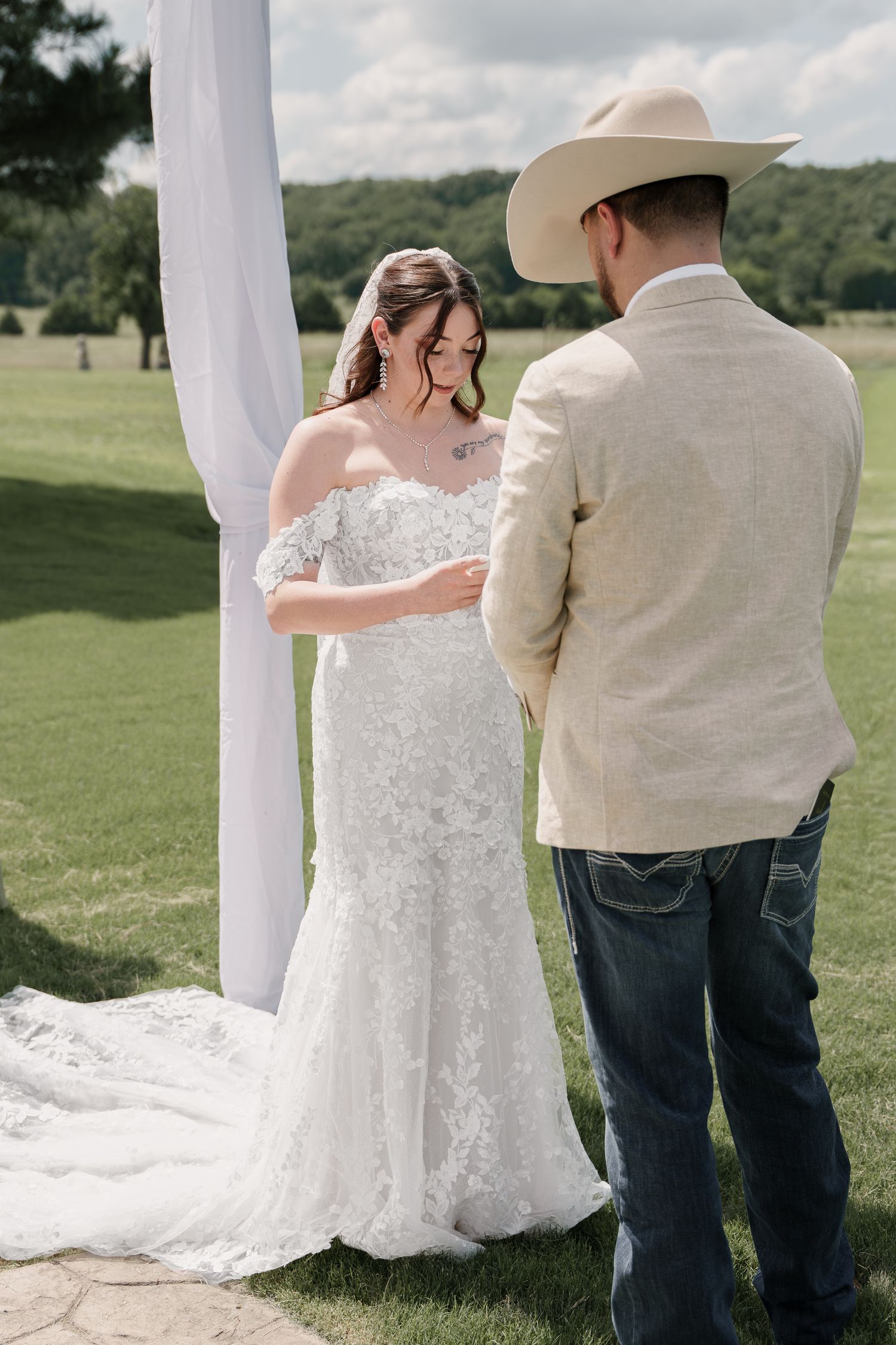Bride in a lace gown faces her groom in a cowboy hat and tan jacket during an outdoor country wedding ceremony.