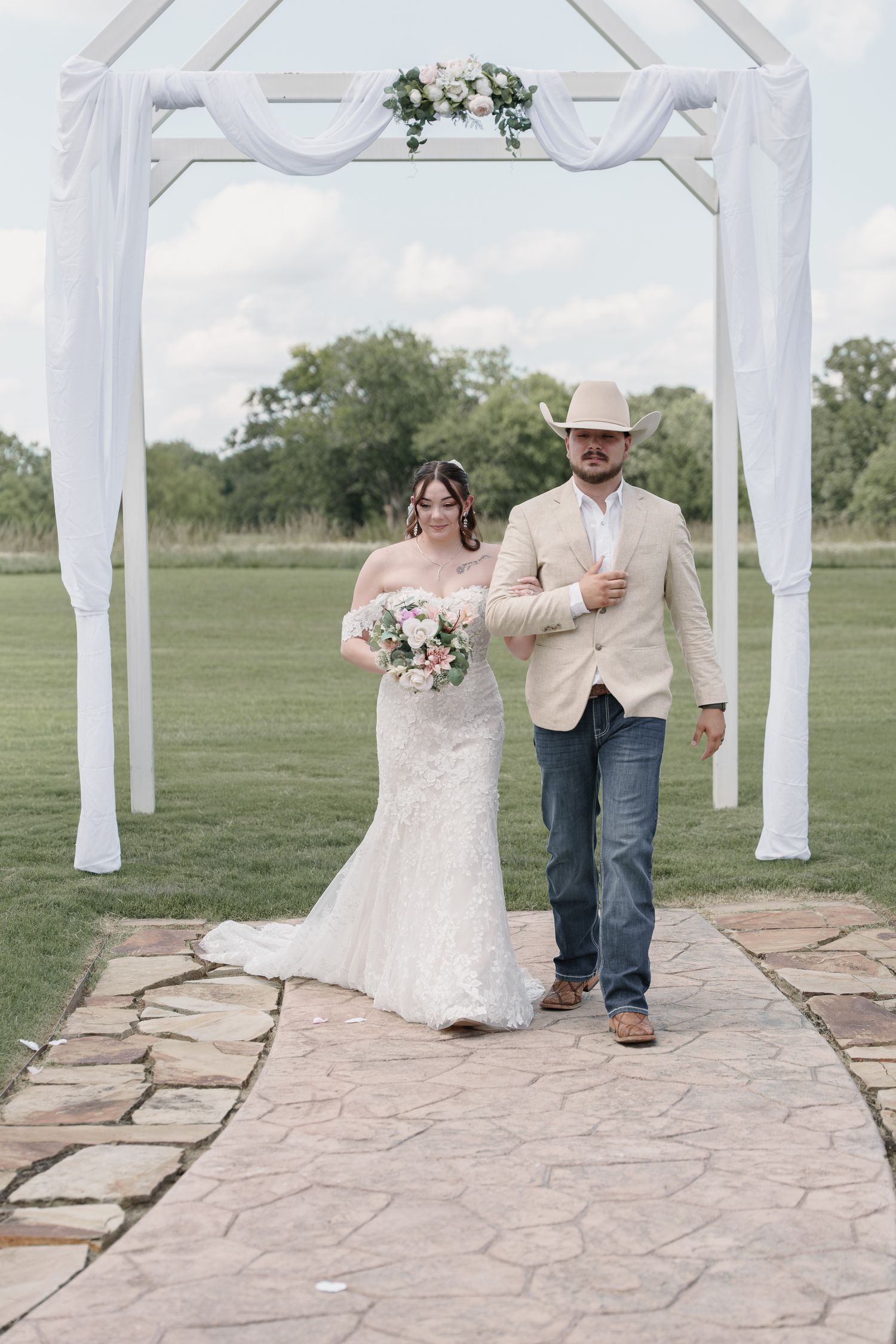 Groom in a cowboy hat escorts the bride in a lace wedding dress down a stone path toward a white floral arch.