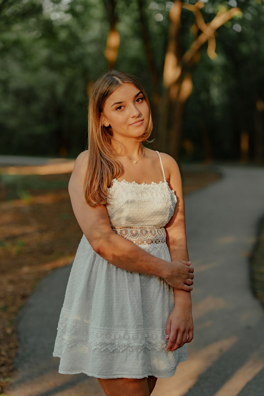 Young woman in a white lace-trim sundress stands on a shaded park path with trees and soft sunlight in the background.