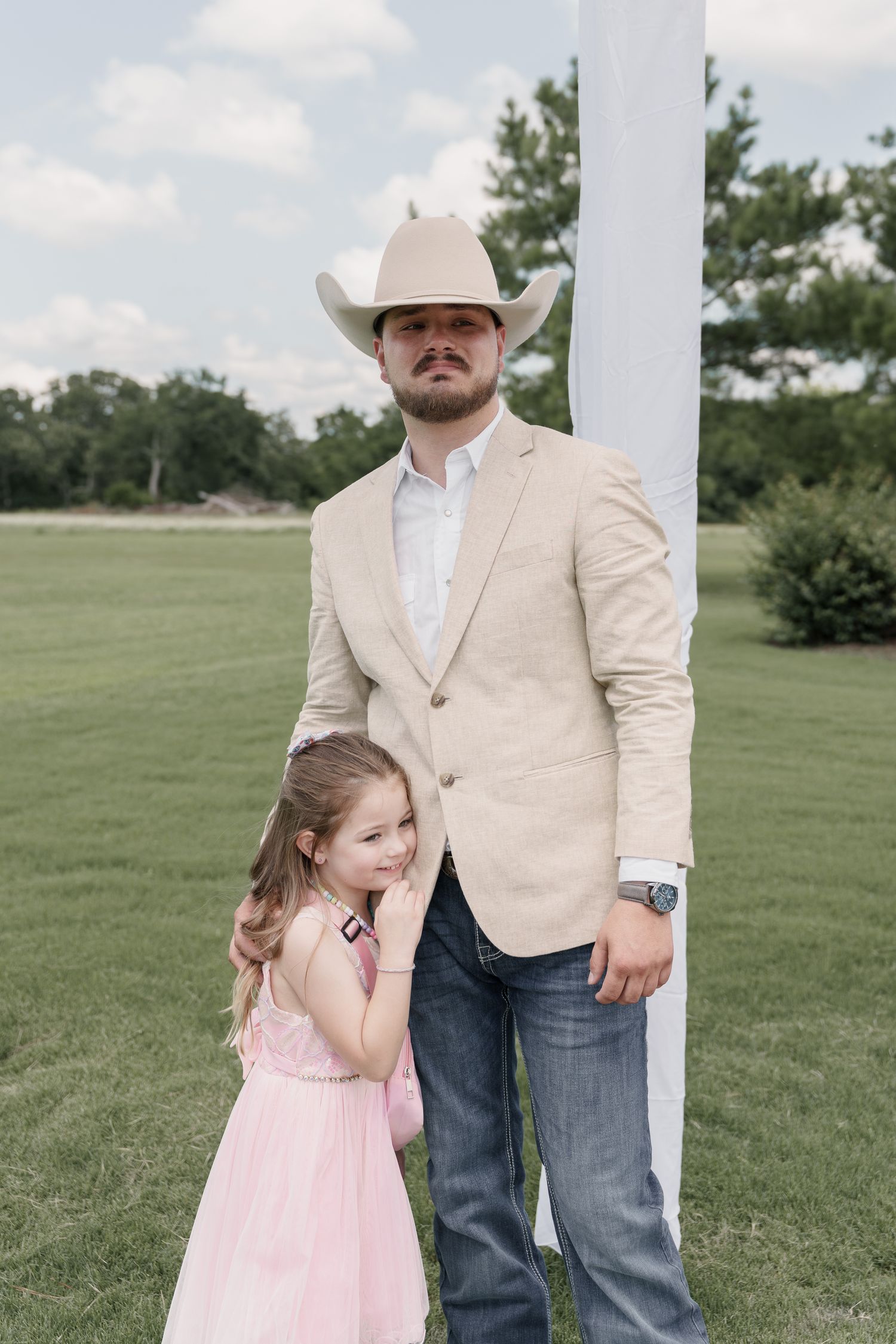 Man in a cream cowboy hat and tan blazer stands on a green lawn as a little girl in a pink dress hugs him at a wedding.