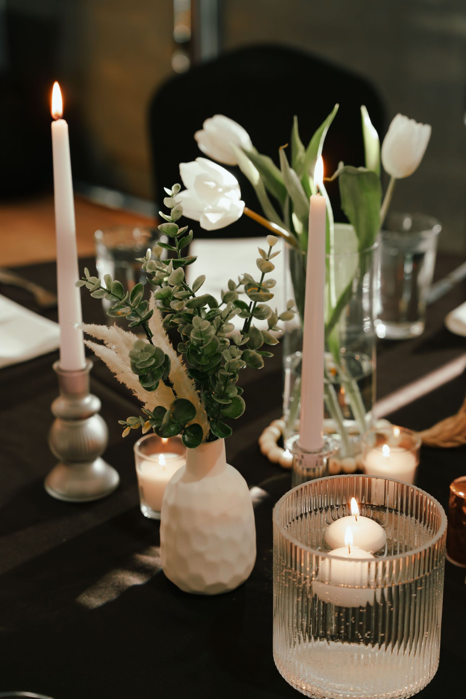 Wedding table centerpiece with white tulips, eucalyptus in a textured vase, tall taper candles and glowing votive candles.